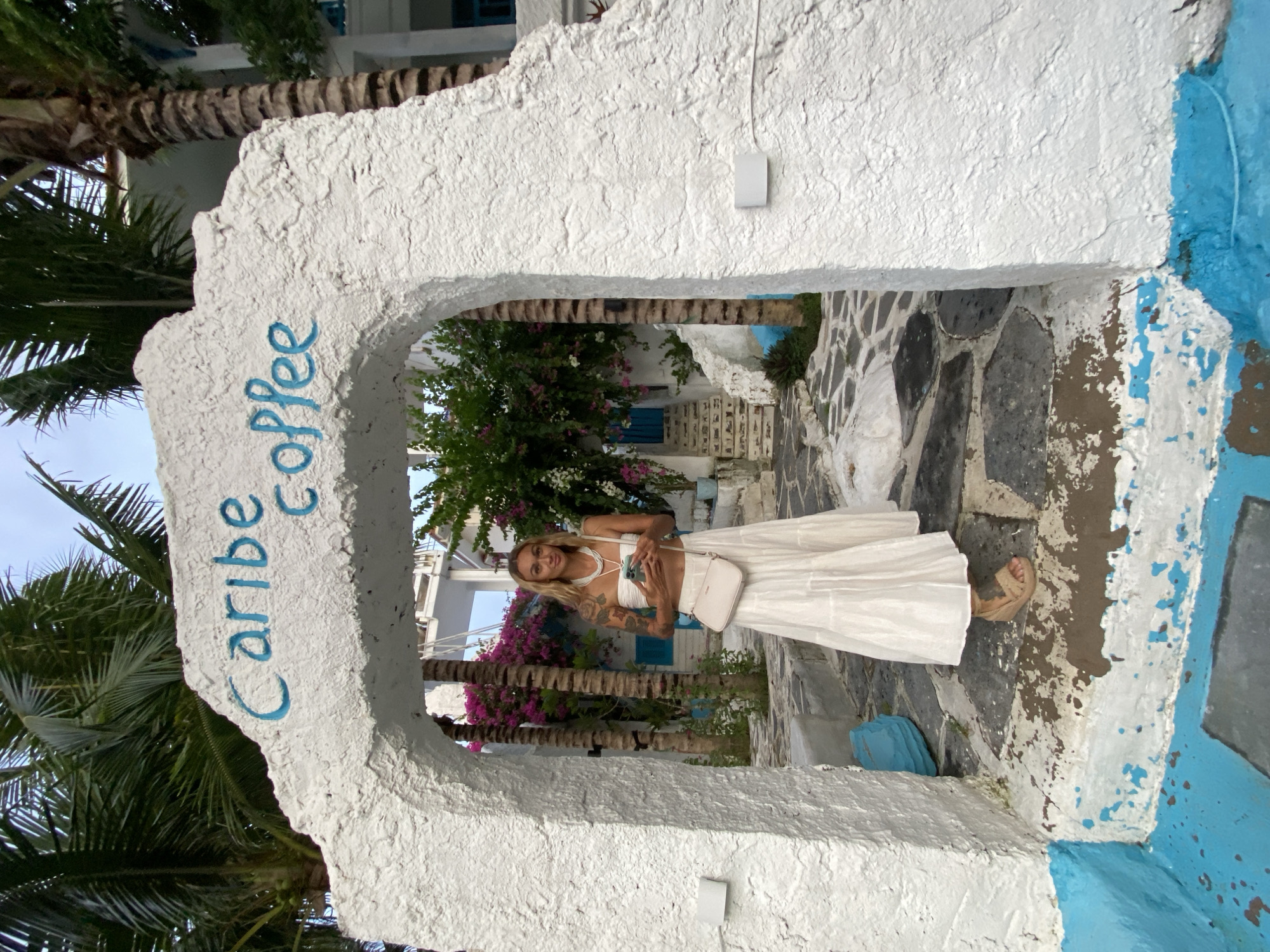 A woman is standing in a white stone archway that reads "Caribe Coffee" in blue lettering, located in Phu Thuy Ward, Vietnam. The archway opens onto a path made of irregular dark stones set in white mortar, leading towards white buildings with blue accents. The woman is wearing a white halter top and a long white skirt, holding a white purse. Palm trees are visible on the left side of the frame, and flowering bushes are visible in the background along the path.