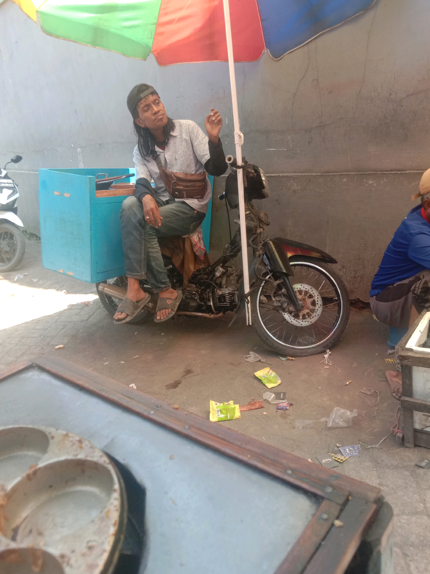 A man wearing a cap, a light-colored shirt, and blue jeans sits on a scooter with a bright blue storage box attached to the back. He has a brown fanny pack around his waist and is wearing dark-colored sandals. The scooter is parked next to the storage box, which has a metal handle visible. A colorful umbrella, with sections of red, green, and yellow, is propped up behind the scooter and the man, providing shade. In the background, a concrete wall appears to be the boundary of the scene. To the right, another person wearing a blue shirt and a cap is partially visible, crouched down. The ground is paved and has scattered debris, including some yellow packaging. The scene suggests an outdoor setting, possibly a street vendor's location in the city of Karang Anyar, Indonesia, given the context.