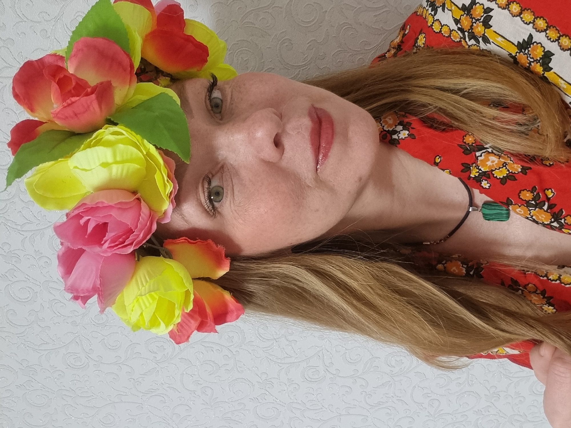 A woman with light skin and freckles wears a flower crown in a close-up, angled shot. The crown is composed of pink, yellow, and red flowers, interspersed with green leaves. She has green eyes and wears lip gloss. Her light brown hair is spread around her head. She wears a red and yellow floral print top and a black necklace with a green, possibly malachite, pendant. The background is a white wall with a subtle, embossed pattern. There is no information available to verify the location of the image as Chaikovsky, Russia.