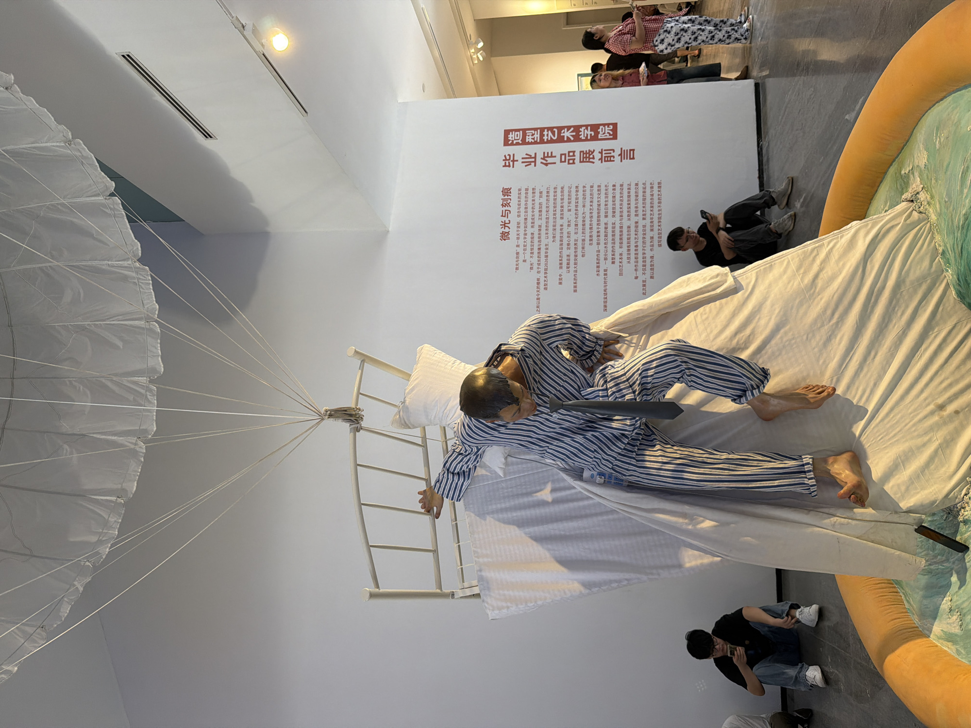 This detailed image captures an indoor art exhibition in Chongqing High-tech Zone, China. The central subject is a sculptural installation featuring a life-sized mannequin and elements resembling a bed. The mannequin, depicting a male figure with short dark hair, is dressed in blue and white striped pajamas and a dark grey tie. Its bare feet are visible, with one foot planted on a white fabric surface and the other slightly raised, suggesting dynamic movement like climbing or falling.
Behind the mannequin stands a white metal bed headboard and footboard frame, oriented vertically. Flowing white fabric, akin to bedsheets, drapes around the bed frame and the mannequin, creating a billowing effect. Above the entire installation, a large white parachute-like canopy is suspended from the ceiling by multiple thin white ropes that converge towards the top of the bed frame.
The base of the installation consists of an orange circular ring, possibly inflatable, encompassing a sculpted turquoise and greenish-blue surface that simulates agitated water with white caps. A dark, rectangular object, possibly a smartphone or small tablet, is partially submerged in this simulated water.
Several people are present in the exhibition space. In the immediate foreground, to the left of the installation, a young East Asian male with glasses, wearing a black t-shirt, blue jeans, and white sneakers, crouches on the dark grey floor, intently looking at a smartphone. Further back and to the right, a small group of visitors stands. One East Asian woman, wearing a red and white checkered shirt and patterned pants, stands with white slippers. Partially visible behind her is another individual in dark attire, and a person with blonde hair in a red top. Another young East Asian male, wearing a black t-shirt and grey pants, is seen sitting on the floor to the left, also engaged with a smartphone.
The exhibition space features plain white walls. Large red Chinese characters on the right wall read "造型艺术学院" (School of Fine Arts / Academy of Plastic Arts) and "毕业作品展前言" (Graduation Exhibition Foreword), with a block of smaller descriptive Chinese text below and to the left of these titles. The ceiling is white, equipped with air vents and recessed lighting. A bright yellow spotlight illuminates the upper right corner. The floor is a dark grey, polished surface.