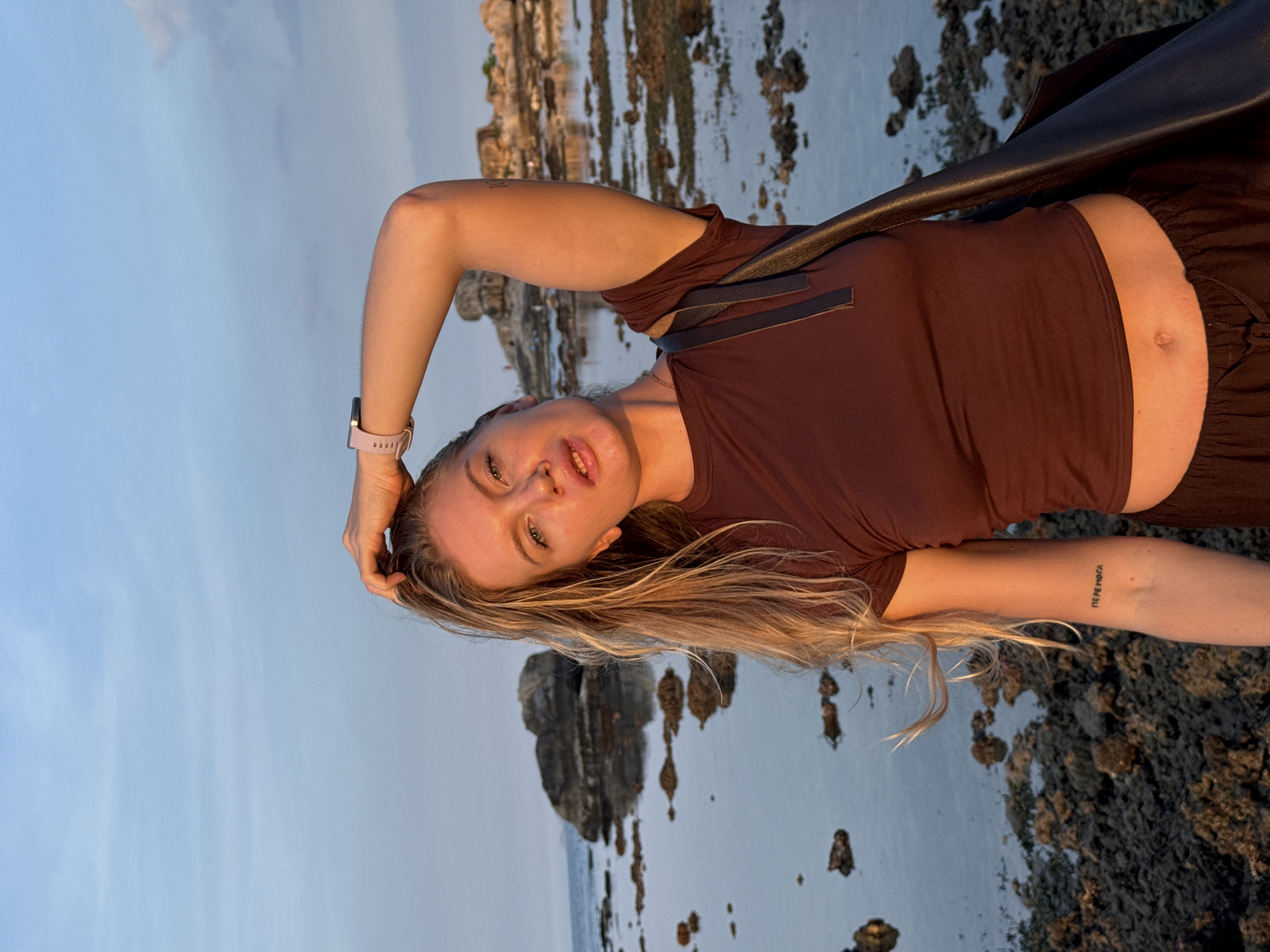 A young woman with blonde hair and a watch on her left wrist is posing for a photo by the sea. She is wearing a brown t-shirt and dark pants. She has a tattoo on her left forearm. In the background, there are rocks and the ocean, with a clear sky above. The image appears to have been taken during the golden hour, as indicated by the warm lighting. The location is not discernible from the image alone.