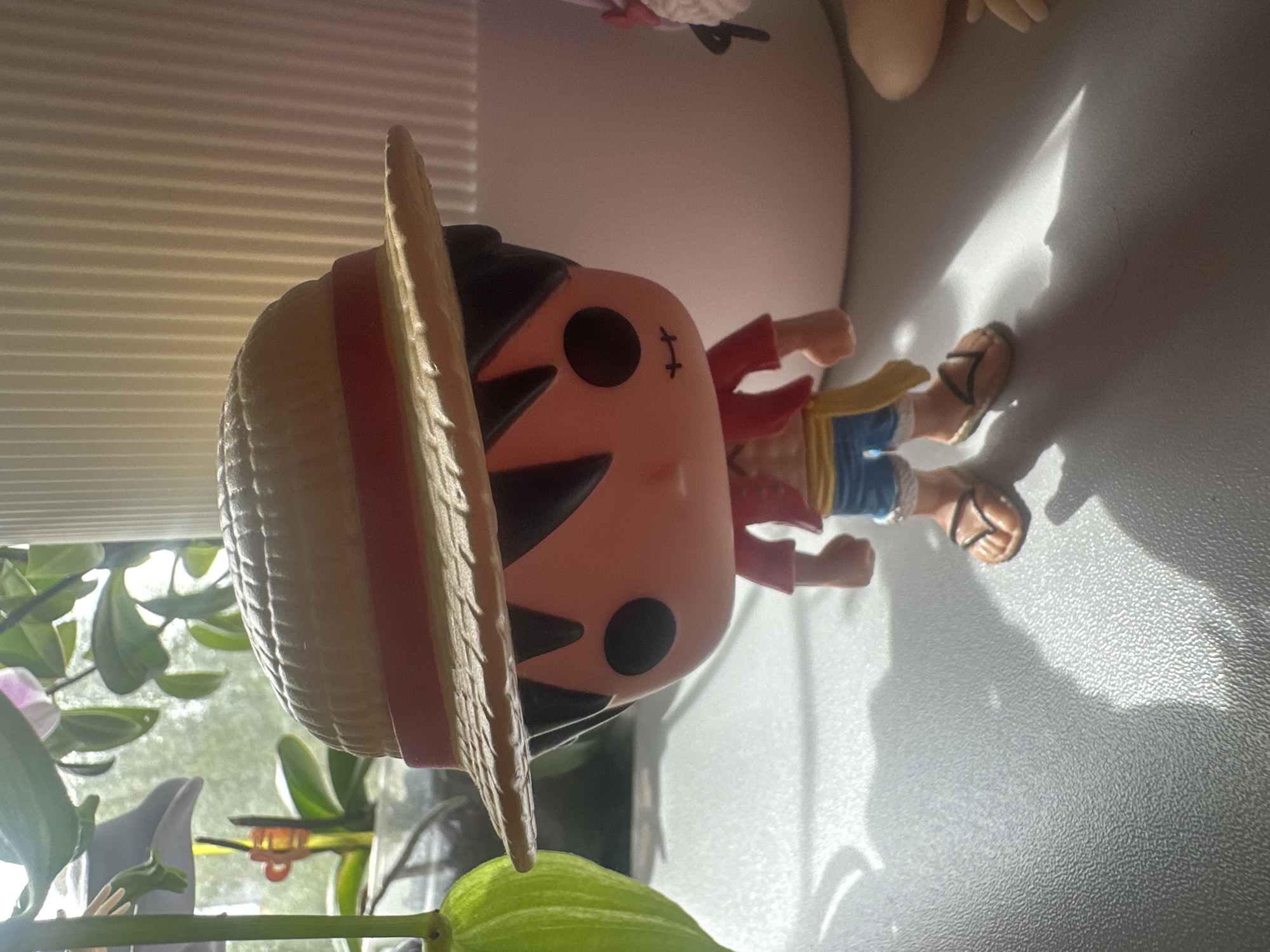 A Funko Pop figurine of Monkey D. Luffy from the anime One Piece is standing on a gray, textured surface. The figurine is wearing its signature straw hat, a red vest over a yellow shirt, blue shorts, and brown sandals. It is positioned in front of a white, ribbed cylindrical object, likely a container or vase. To the left of the figurine, partially visible, is a green plant with broad leaves and a hint of a pink flower. Sunlight is casting shadows on the surface, creating dramatic lighting. The background appears to be a window with diffused light coming through, suggesting an indoor setting near a window. No specific location in Maladzyechna, Belarus, is identifiable from the image.
