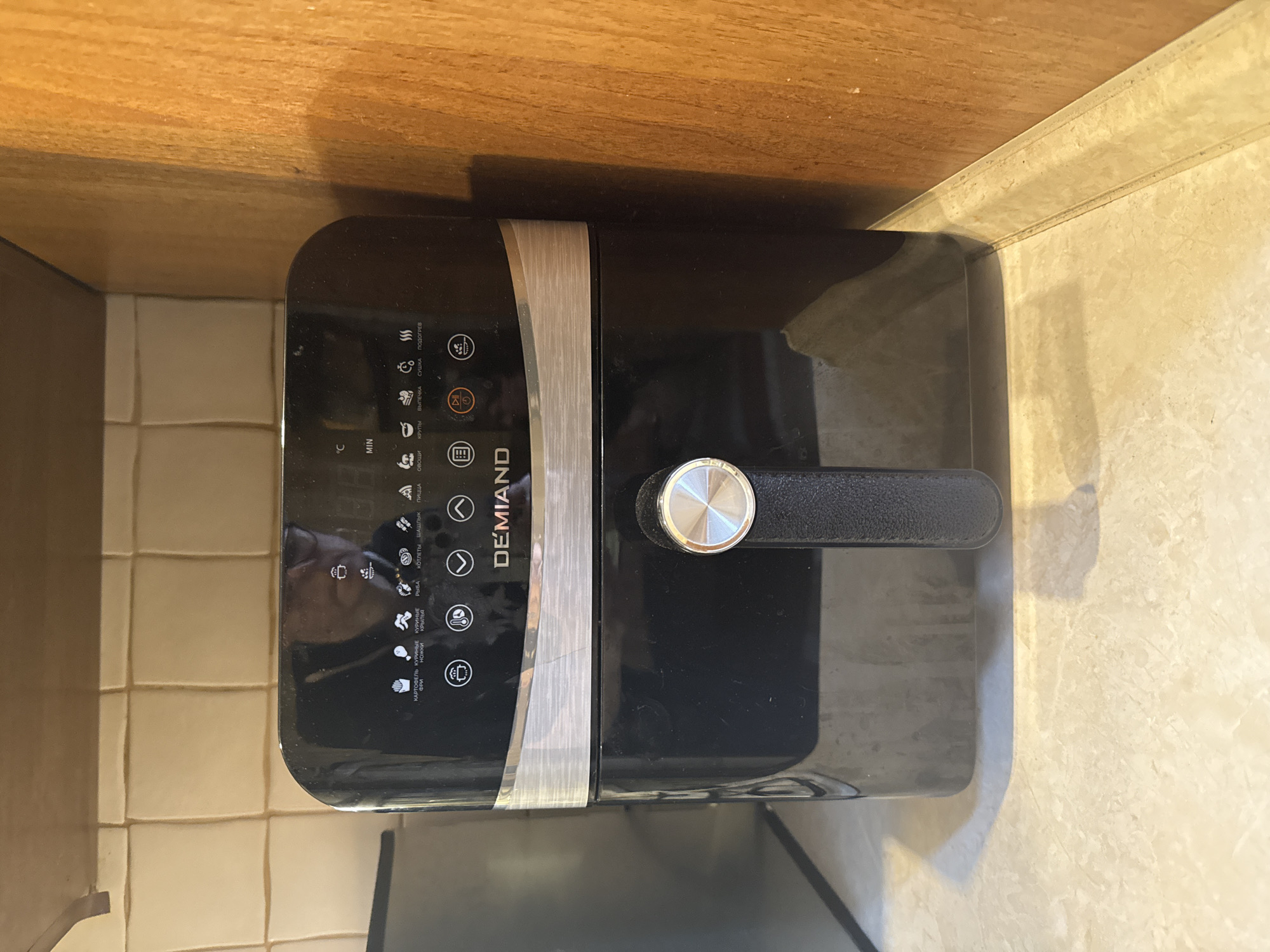 The image shows a black air fryer on a countertop. The air fryer has a silver accent strip and a black handle with a silver dial. The control panel has various icons and text in Russian. The visible text includes "DEMIAND", "MIN", and icons with labels like "КАРТОФЕЛЬ ФРИ" (French fries), "КУРИНЫЕ" (Chicken), "РЫБА" (Fish), "КОТЛЕТЫ" (Cutlets), "ШАШЛЫК" (Shish kebab), "ПИЦЦА" (Pizza), "КРУПЫ" (Grains), "ВЫПЕЧКА" (Baking), "СУШКА" (Drying), and "ПОДОГРЕВ" (Warming). The air fryer is positioned on a beige tiled surface next to a wooden surface, possibly a shelf or cabinet. There is a reflection of a person in the glossy surface of the air fryer. The lighting suggests an indoor setting.