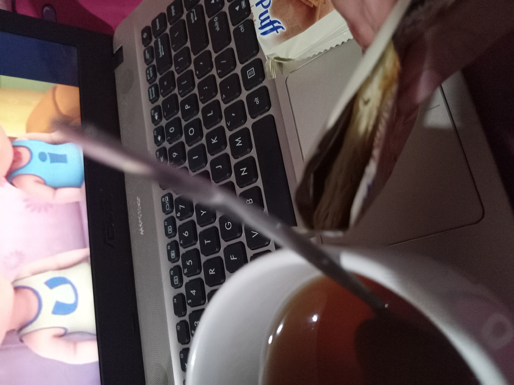 A hand holds open a package of snacks over a laptop keyboard, which is situated next to a white mug of tea. The laptop screen displays an animated scene with two cartoon characters. The overall scene appears to be a domestic setting, likely a living room or bedroom. The image does not contain any information that allows for determining the location in Claket, Indonesia.