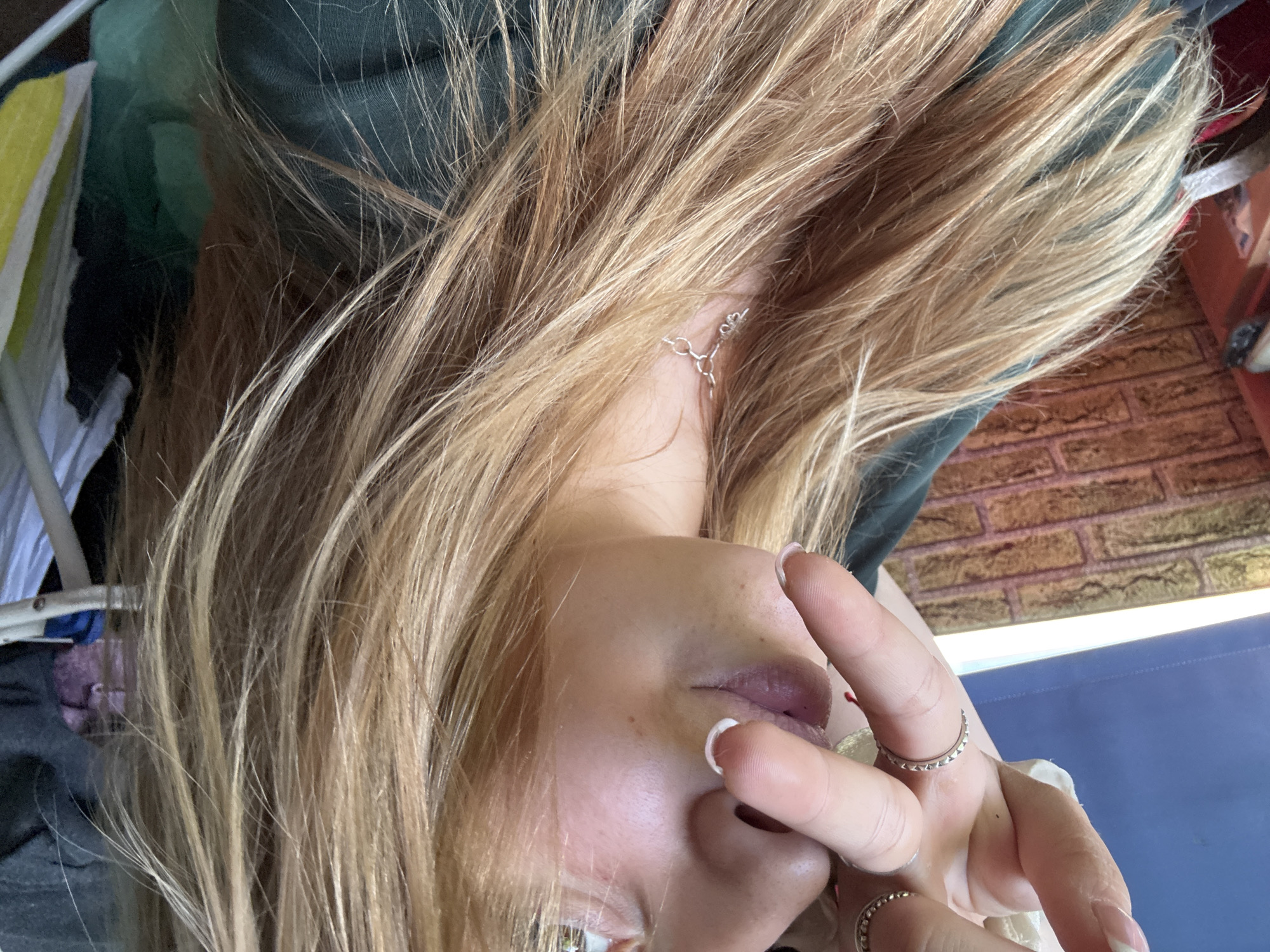 A close-up, slightly rotated image depicts a fair-skinned individual with blond hair and visible silver earrings. The person's face is partially obscured by their own hand, adorned with multiple silver rings. The environment includes a blurred background of green fabric, a section of brick pattern, and a light blue surface. A portion of a white frame structure is visible on the left.