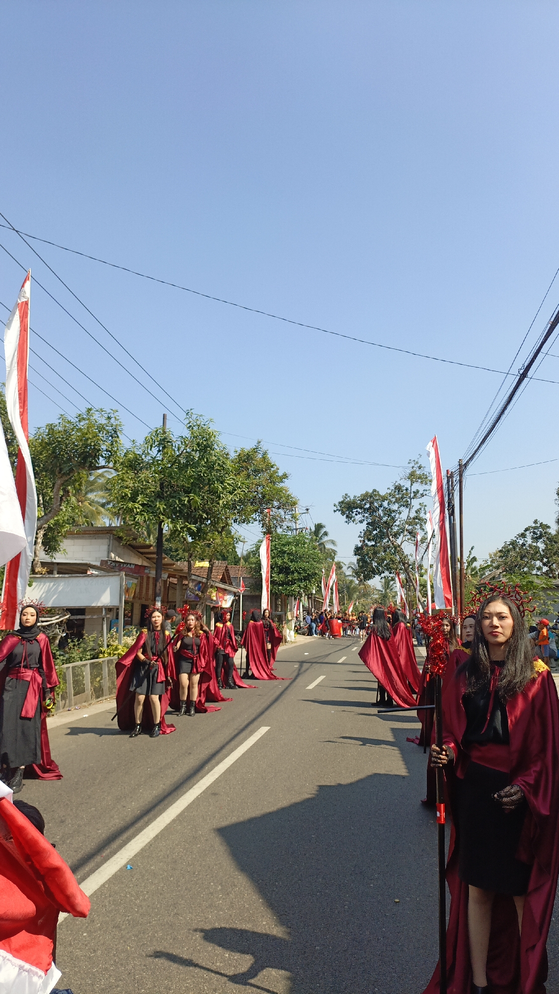 In Krajan Lor, Indonesia, a parade is underway on a street lined with houses and trees. A group of people, primarily women, are participating, dressed in matching crimson cloaks over black outfits. They are adorned with decorative red headpieces, and some are holding flags or staffs. The parade features numerous red and white flags, likely representing the Indonesian flag, displayed on poles along the roadside. The sky is clear and blue, indicating a sunny day. The street is paved, with white lines marking the lanes. The participants are walking in formation, suggesting a performance or procession. In the background, a few spectators can be seen observing the event. Overhead, power lines crisscross the sky.
