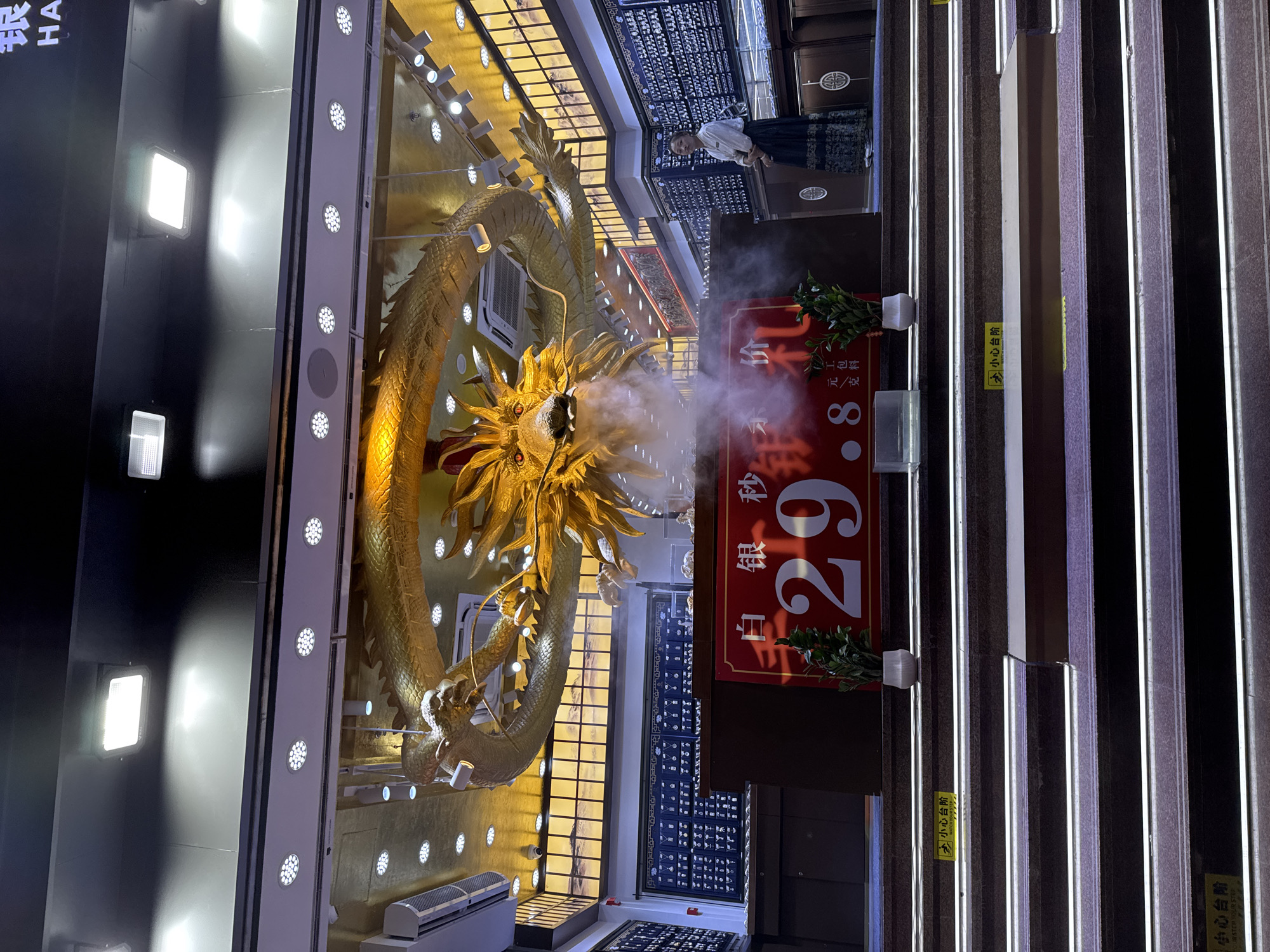 The image shows a store interior, likely a jewelry store, in Shanghai, China. A large, golden dragon sculpture hangs from the ceiling, appearing to breathe mist. In the foreground, a red sign displays large white numbers "29.8", suggesting a price or a date. A woman in traditional attire stands to the right, looking towards the viewer. The store appears to be well-lit with multiple spotlights and illuminated display shelves filled with items. The overall atmosphere suggests a commercial establishment with a focus on traditional Chinese aesthetics.