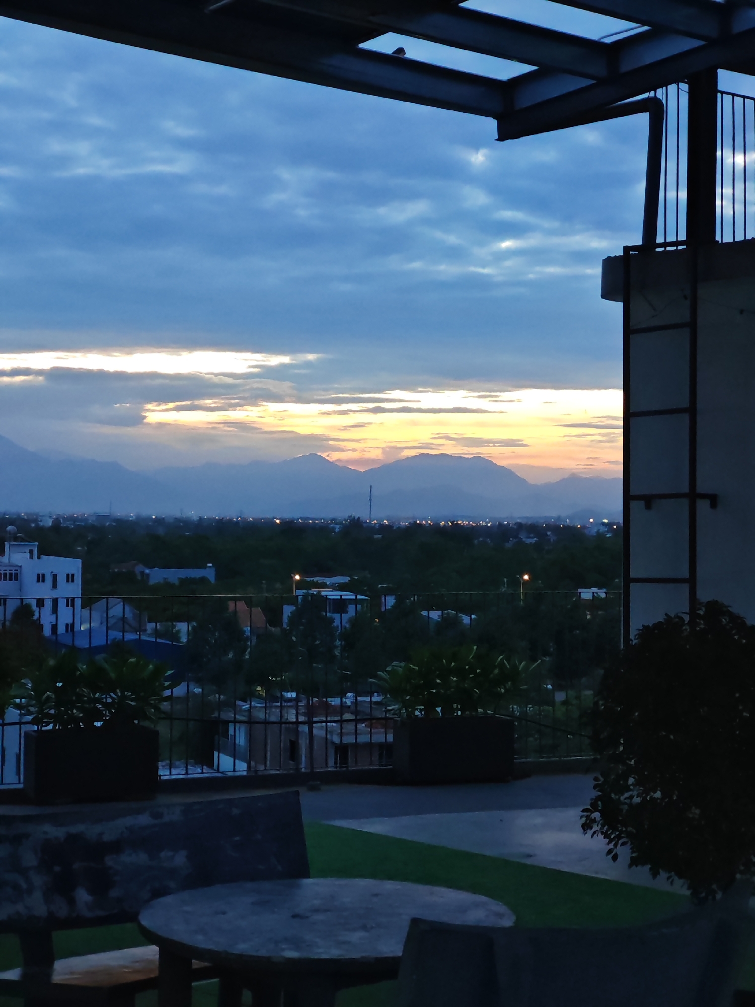The image depicts a twilight view from a rooftop in Da Nang, Vietnam. In the foreground, a concrete table and bench are visible on a paved area with patches of artificial turf. Beyond the seating, planters with green foliage are placed along a metal railing. The middle ground shows a cityscape with buildings, trees, and streetlights, extending towards a range of silhouetted mountains in the distance. The sky is filled with dramatic clouds, illuminated by the fading light of sunset, casting a warm glow on the horizon. The architectural elements of the building, including a metal overhang and vertical support structures, frame the expansive view.