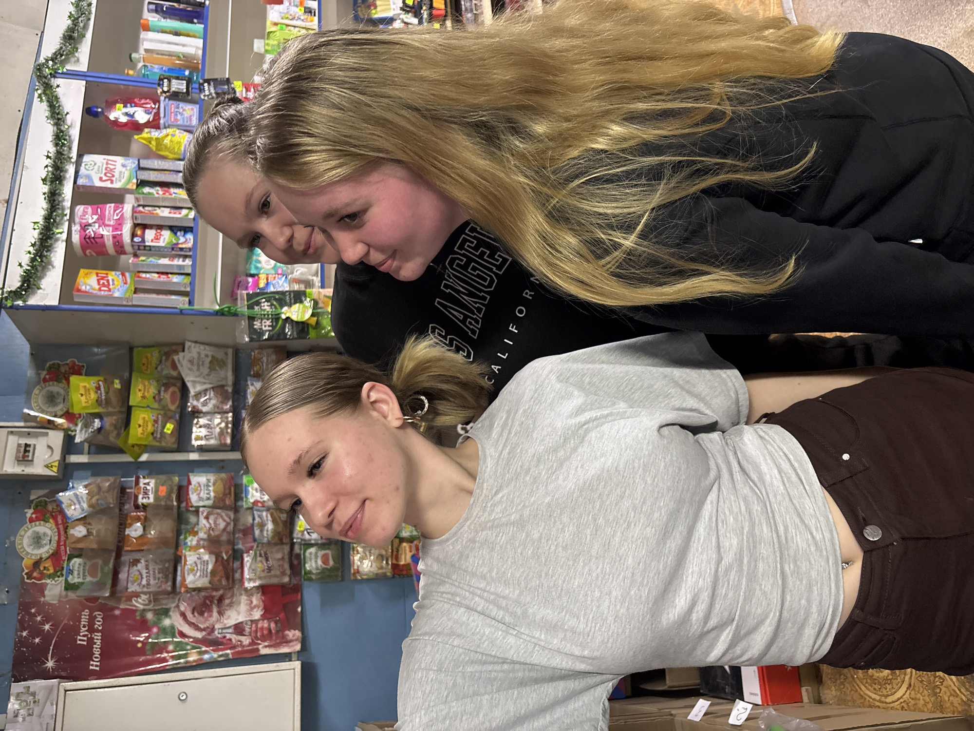 Three young women are browsing the shelves in a small store. Two of the women are fair-skinned, with long blonde hair; one wears a black shirt with “LOS ANGELES CALIFORNIA” printed on the front. The third woman has fair skin, and her hair is pulled back into a ponytail; she is wearing a grey t-shirt and brown pants. The shelves are lined with packages of food, toilet paper, and various household items. A poster with a New Year's greeting in Russian, “Пусть Новый год”, is affixed to the side of the shelving unit. The overall appearance of the store suggests a modest, possibly rural, setting.