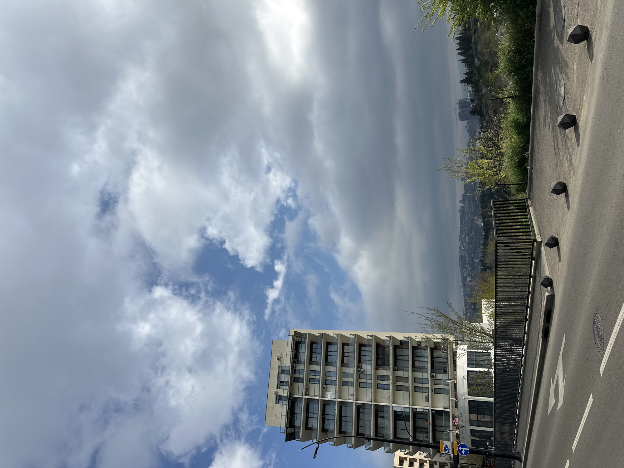 The image shows a view in Tbilisi, Georgia, featuring a modern, multi-story building with balconies on each floor. The building appears to be of mid-century architectural style. The sky above is filled with a mix of light and dark clouds. A street runs along the right side of the frame, bordered by a black metal fence. Geometric, black bollards are placed along the edge of the road. Beyond the fence and across the street, there is a view of the city and distant hills partially obscured by overcast skies and haze. A few trees and greenery are interspersed in the mid-ground. The image is taken from a slightly elevated position.
