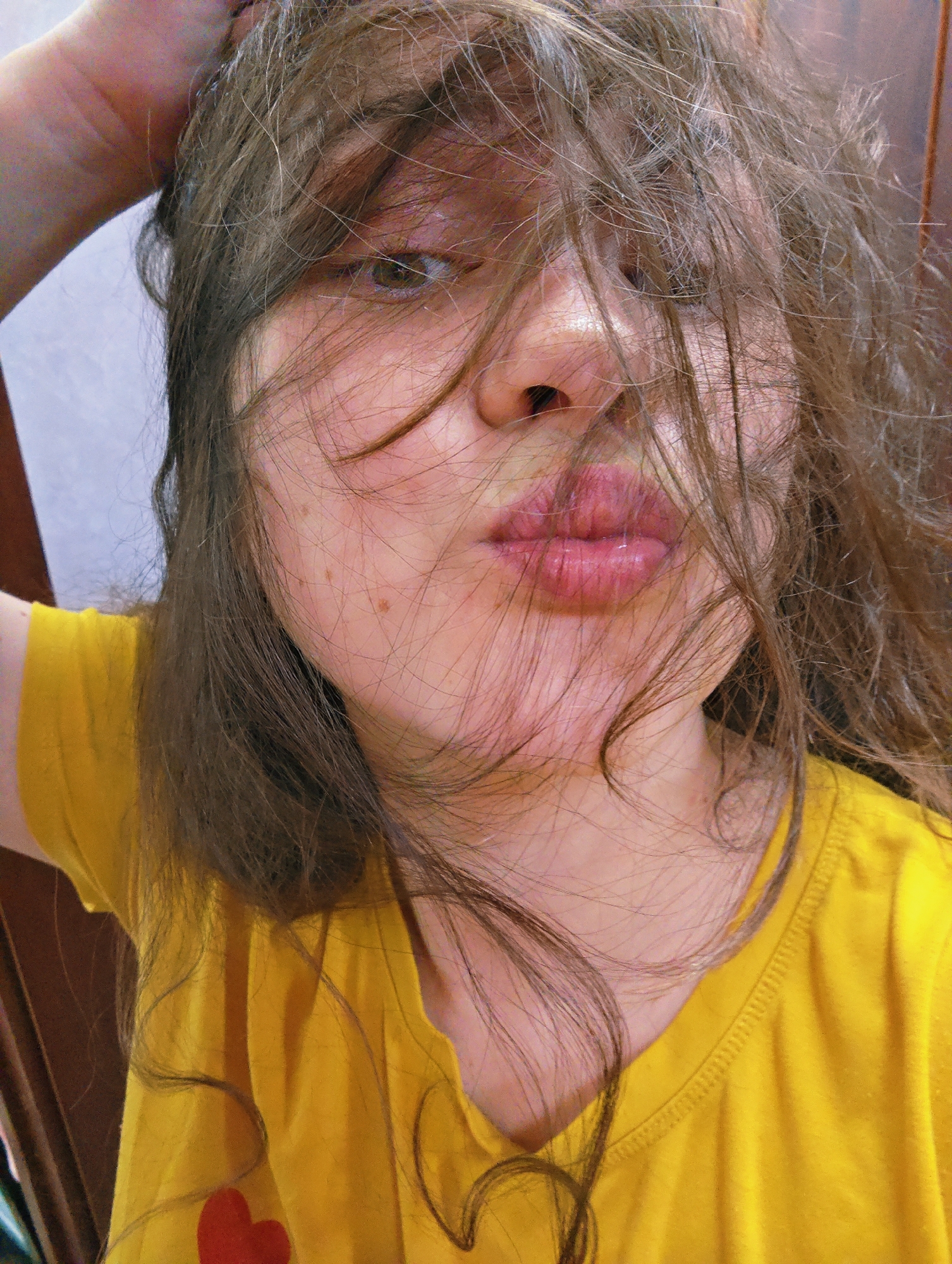 A selfie of a fair-skinned person wearing a yellow t-shirt with a red heart emblem on the lower left. Their brown hair is disheveled and partially obscures their face. They are puckering their lips in a kissing gesture, and their left arm is raised with their hand touching their head. The background is a light-colored wall on the left and a dark wooden door on the right. The location information is not discernible from the image.
