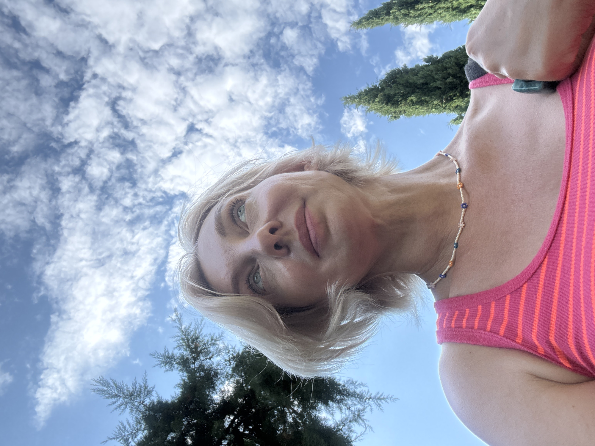 An outdoor, low-angle, medium shot captures a woman with short, blonde hair and fair skin looking up at the sky. She is wearing a pink and orange striped tank top and a beaded necklace. Her right hand is raised and holding a dark green object. Cypress trees and a bright blue sky with white clouds form the background. The scene appears to be located in Sochi, Russia, given the distinctive cypress trees and coastal climate often associated with the city.