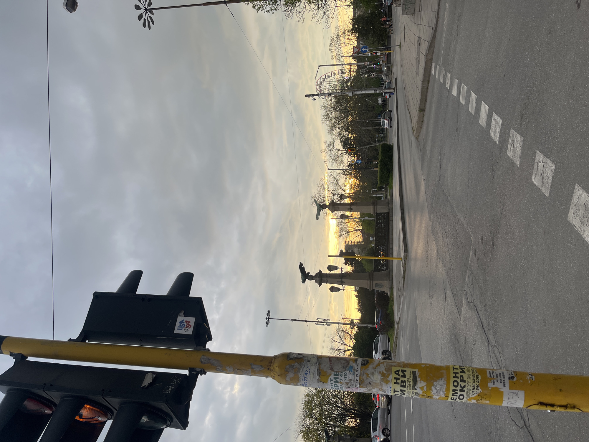 The image captures an urban street scene in Sofia, Bulgaria, oriented vertically (requiring a 90-degree clockwise rotation for proper viewing). The time of day appears to be either sunset or sunrise, as indicated by the low, golden sunlight breaking through clouds and casting a warm glow across the sky and distant elements.
In the immediate foreground, a yellow traffic light pole dominates the left side of the frame (when viewed upright). This pole is heavily covered with weathered paper advertisements and stickers, many of which feature Cyrillic text. One notable sticker on the black traffic light housing itself shows "USA 40" alongside a small football icon. The traffic lights are black, and one displays an orange light.
Looking across the street, a road with dashed white lane markings is visible. Several cars are present, both driving and parked along the street. Beyond the road, a green area with trees and shrubs borders a distant horizon. Prominently silhouetted against the bright sky are two monument-like structures, each featuring a large bird figure, possibly an eagle. Further in the background, a large Ferris wheel is visible above the tree line, suggesting a nearby park or amusement area. The sky above is mostly cloudy, with the sun's warm light concentrated near the horizon.
Visible Cyrillic text on the traffic light pole includes phrases like "ОТ НАС КАЧЕСТВО" (From us, quality), "ОТ ВАС ДОВЕРИЕ" (From you, trust), and partial words related to "Ремонт" (Repair) and "услуги" (services), along with various phone numbers such as "0876 77 91 40" and "0893 207 2680". These texts indicate local advertisements for different services. The overall scene depicts a typical urban environment during a transitional light period.