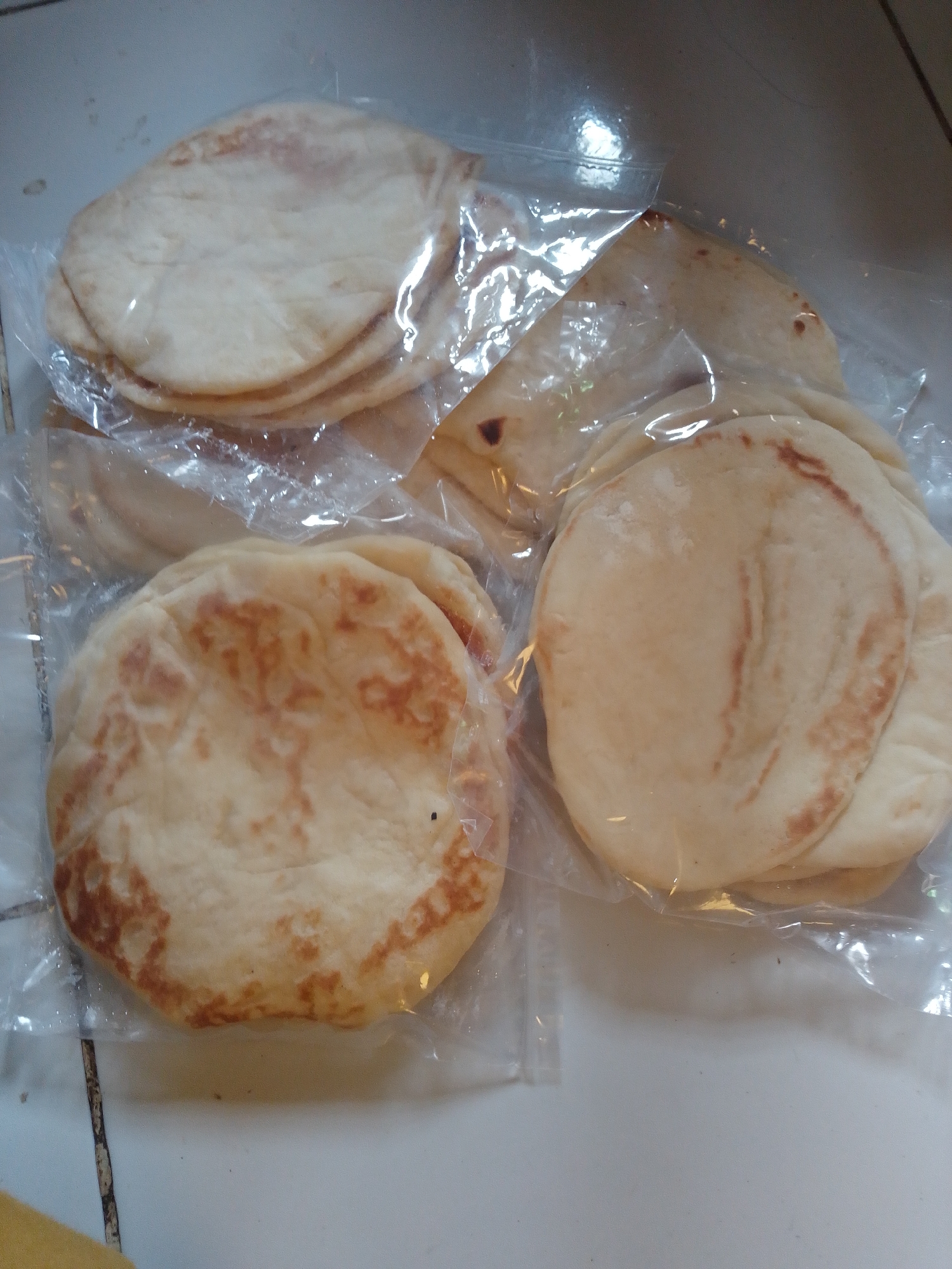 The image shows a top-down view of several packages of flatbread, likely pita bread, on a tiled surface. The flatbread is light beige with some brown char marks. The packages are clear plastic, and the flatbread is stacked within them. The background is a plain, light-colored tiled surface with visible grout lines. The lighting is somewhat dim and casts a slight sheen on the plastic packaging. There are no people or identifiable landmarks in the image. The location in Panyingkiran, Indonesia, is not visually represented in the image itself, but is provided as context.