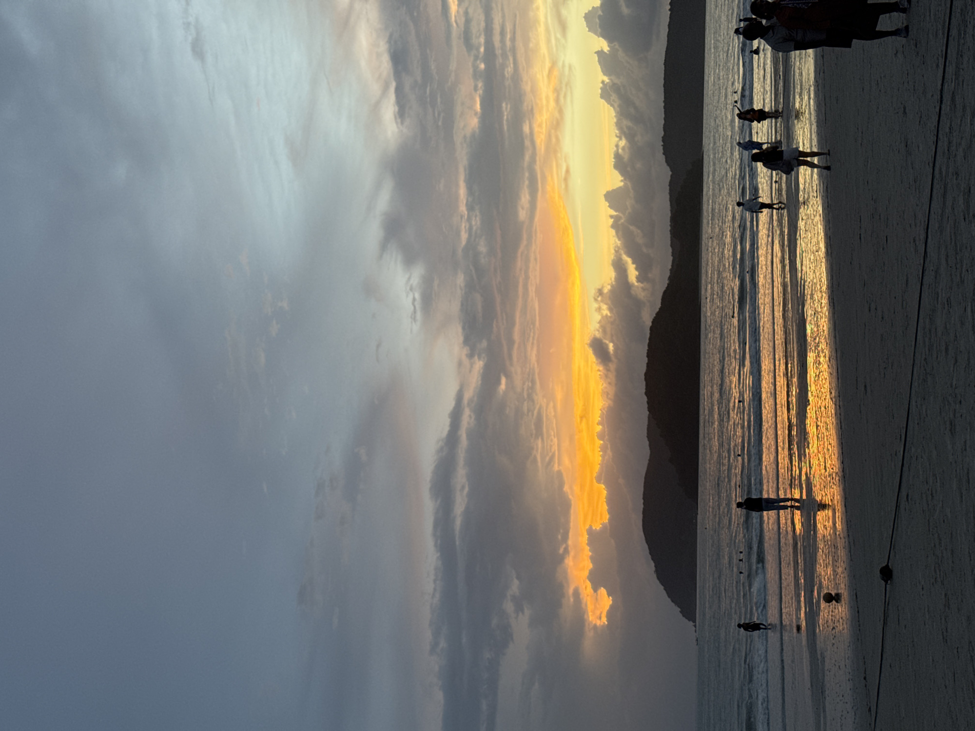 The image depicts a sunset scene at Cenang Beach, Malaysia. Silhouetted figures of people are visible on the wet sand near the water's edge. Some individuals are walking along the beach, while others are in the shallow water. The sun is setting, casting a vibrant orange and yellow glow on the clouds and reflecting on the ocean's surface. The sky is a mix of blue and grey clouds, with the dramatic colors of the sunset concentrated towards the horizon. Dark, undulating hills form the backdrop across the water. The foreground shows the sandy beach with a dark rope and buoys.