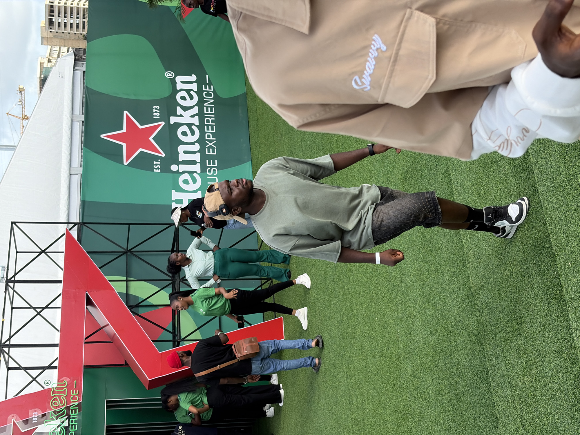 The image depicts a group of people at what appears to be a Heineken promotional event, possibly in Lagos, Nigeria. A large Heineken logo is visible in the background, along with a partially visible building in the distance and a construction crane. Several individuals are present, including one man prominently positioned in the foreground wearing a khaki jacket with the word "Swarvy" embroidered on it. A man wearing a green shirt, brown cap, and headphones is slightly further back. In the background, other people are standing or walking, some wearing green shirts. The ground is covered with artificial grass.