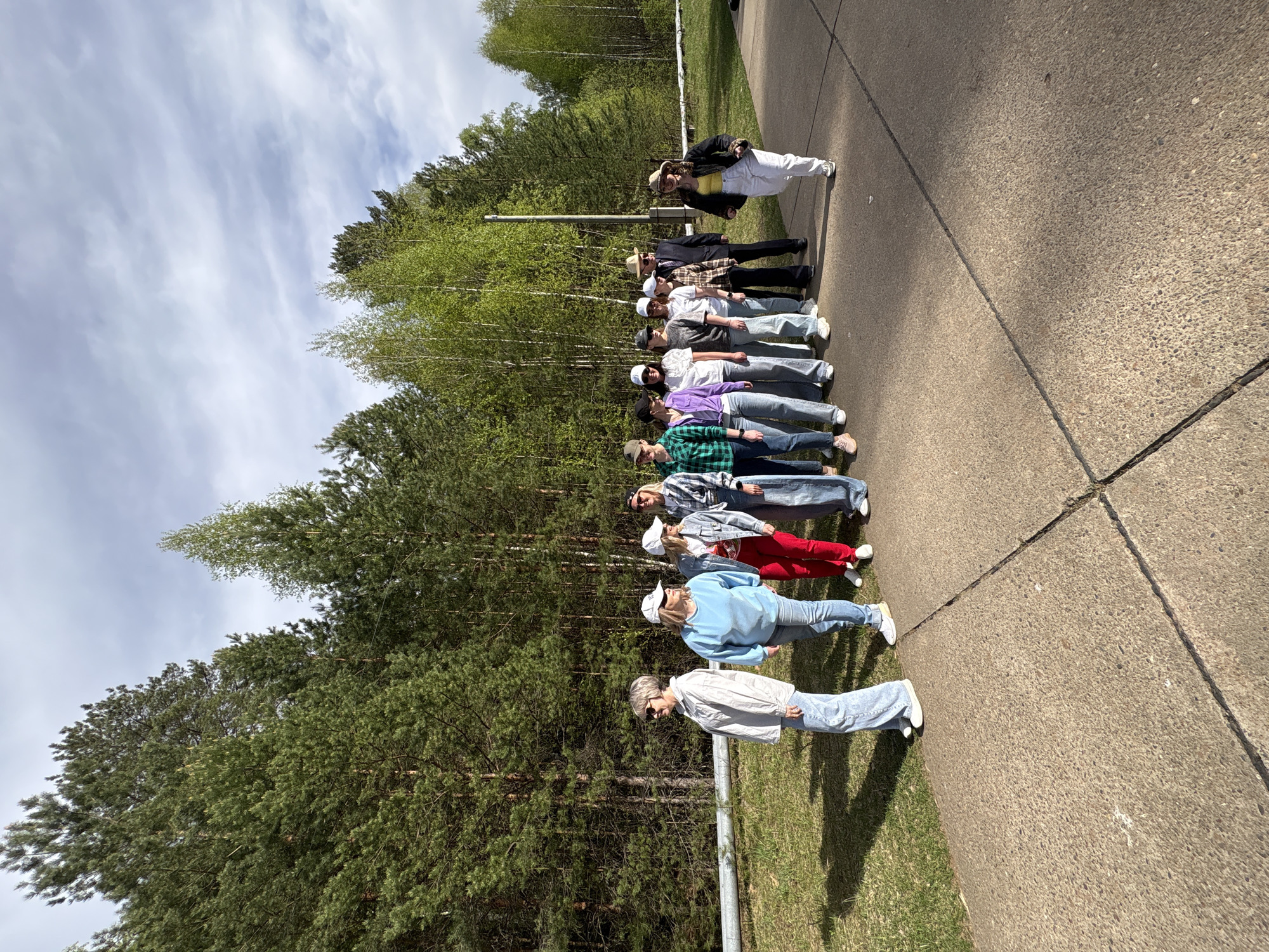 A photograph taken in Марковский, Russia, depicts a group of twelve individuals standing in a line on a concrete walkway adjacent to a wooded area. The individuals are of varying ages and dressed in casual clothing. The walkway appears to be part of a park or recreational area, with trees visible in the background. The sky is partly cloudy. There are no visible buildings or structures, indicating the scene is in a natural or semi-natural environment within the city.