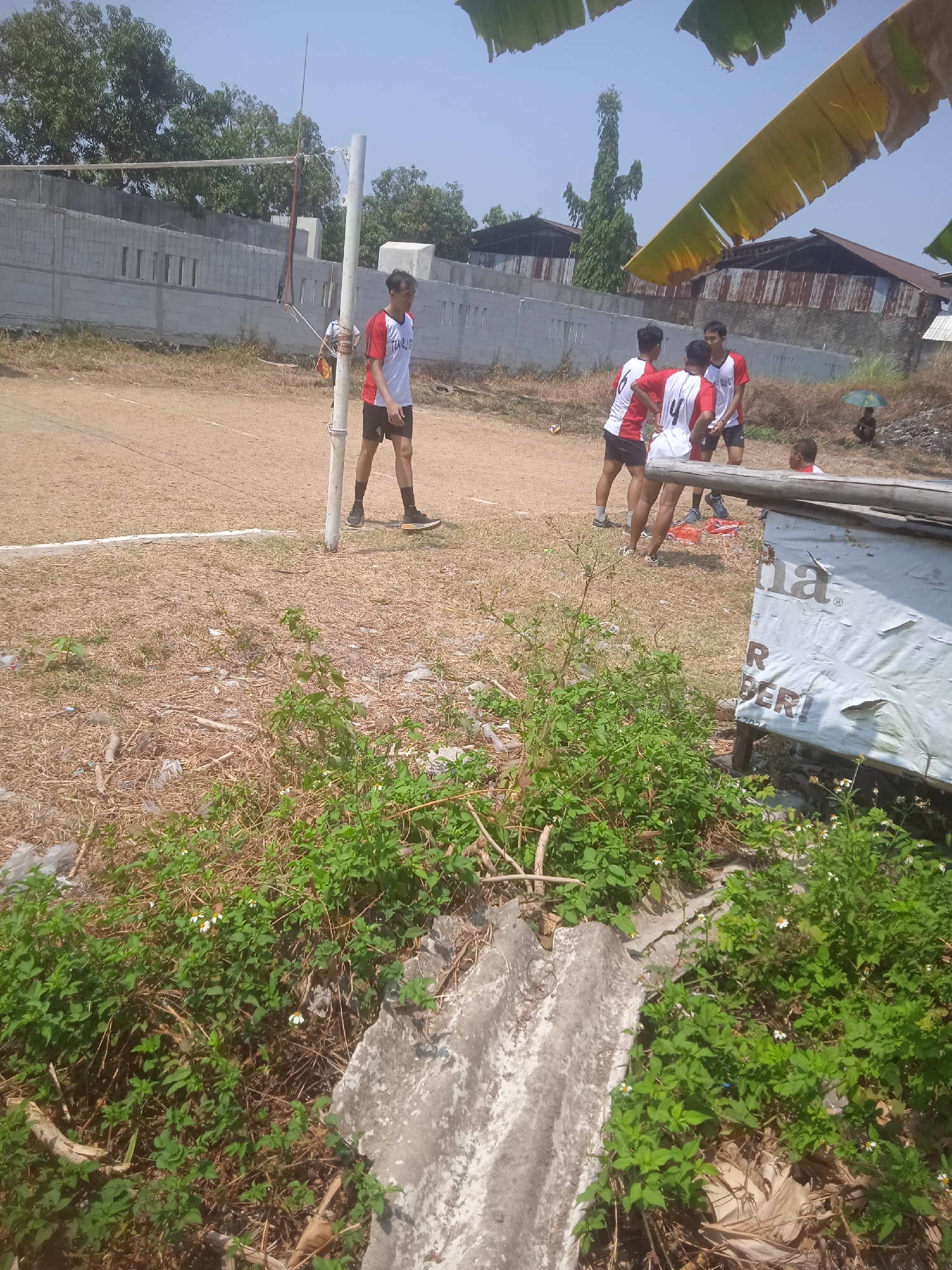 This image depicts a group of young men engaged in a volleyball game on a dirt court, likely located in Klodran, Indonesia. The court is marked with white lines, and a volleyball net is set up, though its tension appears slack. The players are wearing athletic attire, predominantly red and white jerseys and dark shorts. One player in the foreground stands near a white volleyball pole. In the background, a light-colored concrete wall with rectangular openings runs along the property line. Beyond the wall, several buildings with corrugated metal roofs are visible, suggesting a semi-urban or rural setting. Some vegetation, including trees and dried grass, surrounds the court. In the lower foreground, lush green plants and some debris, including broken concrete and plastic, are present. A partially covered wooden structure with text printed on it is also visible on the right side of the image. The sky is clear and bright blue, indicating a sunny day.