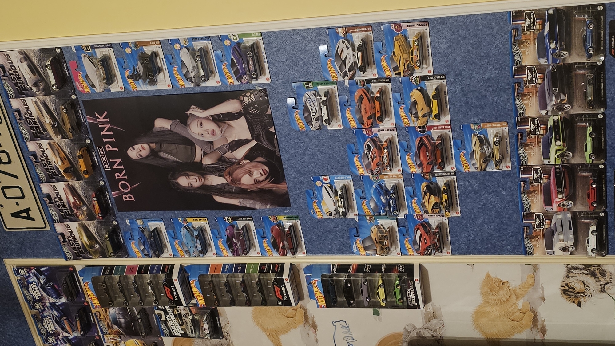 The image shows a collection of Hot Wheels toy cars displayed on a blue textured surface. The cars are mostly in their original packaging. A poster of the K-pop group BLACKPINK, specifically their album "Born Pink", is positioned in the center. To the left is a license plate with "A-078" visible. A shelf-like structure below the display features more toy cars in packaging, and a patterned surface with illustrations of cats. The location, Varna, Bulgaria, is not discernible from the image alone.