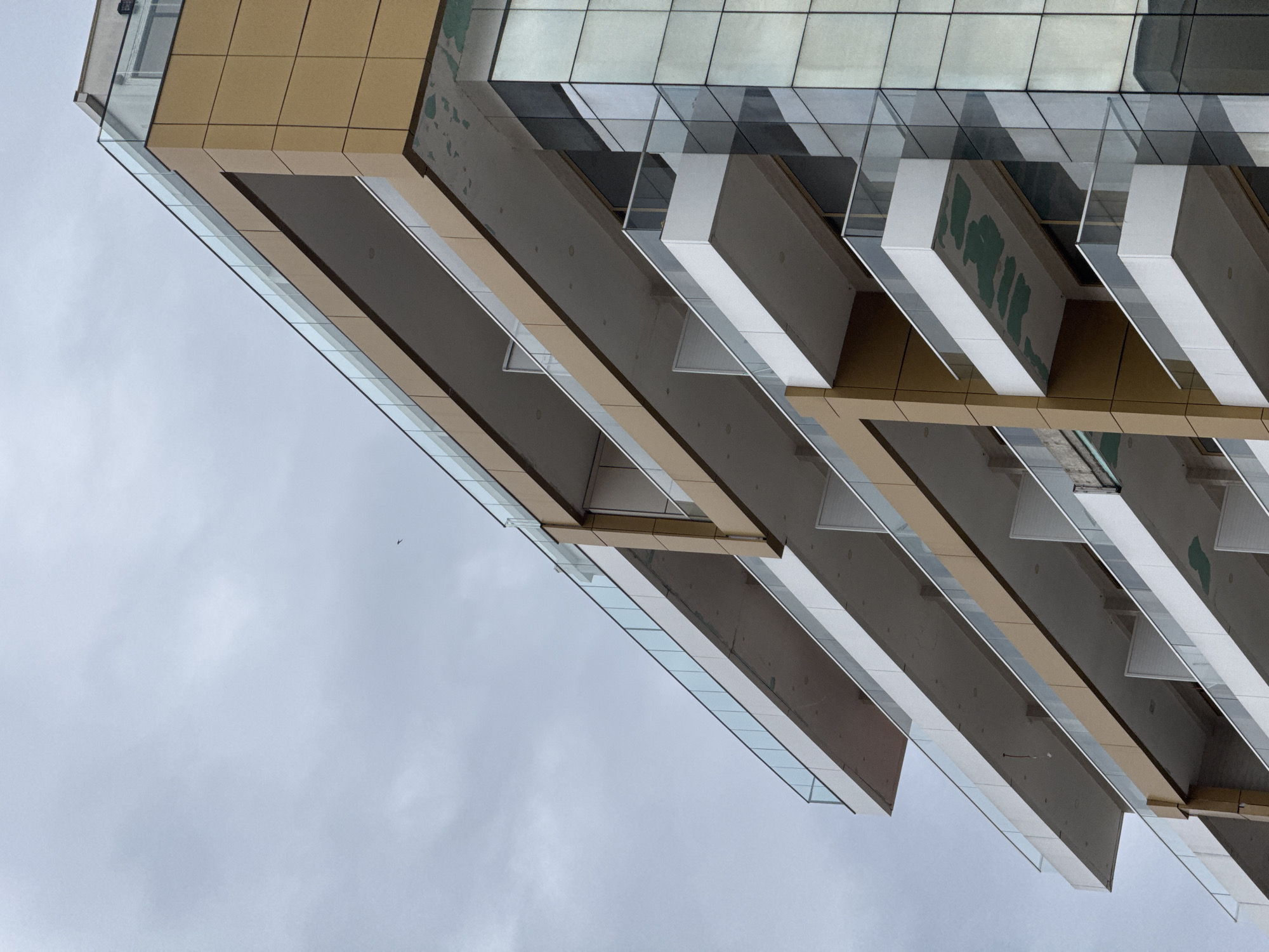 This upward-angled photograph captures the architectural details of a modern building against an overcast sky. The structure features a complex interplay of geometric forms and materials. The upper sections of the building are clad in light brown/tan rectangular panels and large reflective glass windows, which show subtle distortions and reflections of the environment.
Below these sections, multiple parallel layers of balconies or cantilevered floors extend outwards. These are characterized by smooth, dark grey undersides and distinctive white, angled panels forming the edges and supports. Clear glass balustrades run along the outer perimeter of these structures, adding to the contemporary design.
The perspective is from a low vantage point, creating strong diagonal lines that dominate the composition. The sky, visible in the lower left portion of the image, is uniformly grey and cloudy, indicating an overcast day. A very small, distant bird is barely visible as a dark 'V' shape in the sky on the left side. There is no discernible human activity or legible text. The image highlights the clean lines and modern aesthetic of the building, typical of developments found in locations like Budva, Montenegro.