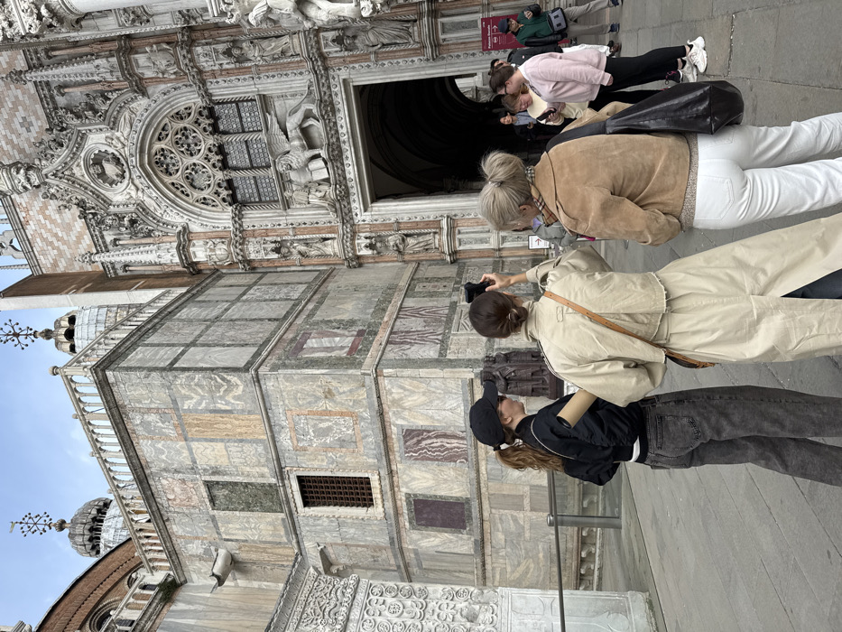 An outdoor scene in Venice, Italy, captures a group of individuals observing the intricate architecture of the Doge's Palace. The prominent building on the right features a grand Gothic entrance, identified as the Porta della Carta, adorned with elaborate carvings, numerous statues within niches, and a central relief sculpture of a winged lion holding a book above a large arched doorway. To the left of this entrance, an adjoining section of the palace facade is constructed from alternating light and dark marble panels arranged in geometric patterns, interspersed with barred windows and topped by a decorative balustrade. Domes with distinctive cross-shaped finials are visible against the overcast sky in the upper background, indicative of St. Mark's Basilica's proximity.
In the foreground, three people are positioned with their backs or sides mostly towards the viewer. On the left, a person wearing a black baseball cap, black bomber jacket, and dark grey jeans holds a rolled document or scroll. In the center, a person wearing a long beige trench coat and a brown shoulder bag holds a black smartphone horizontally, actively photographing or recording the architectural details. To their right, a third individual, with blonde hair styled in a bun, wears a light brown suede jacket, white jeans, and carries a black shoulder bag. Further in the mid-ground and background, additional people are present, either standing or walking, near other entrances to the building complex. The ground surface is paved stone.