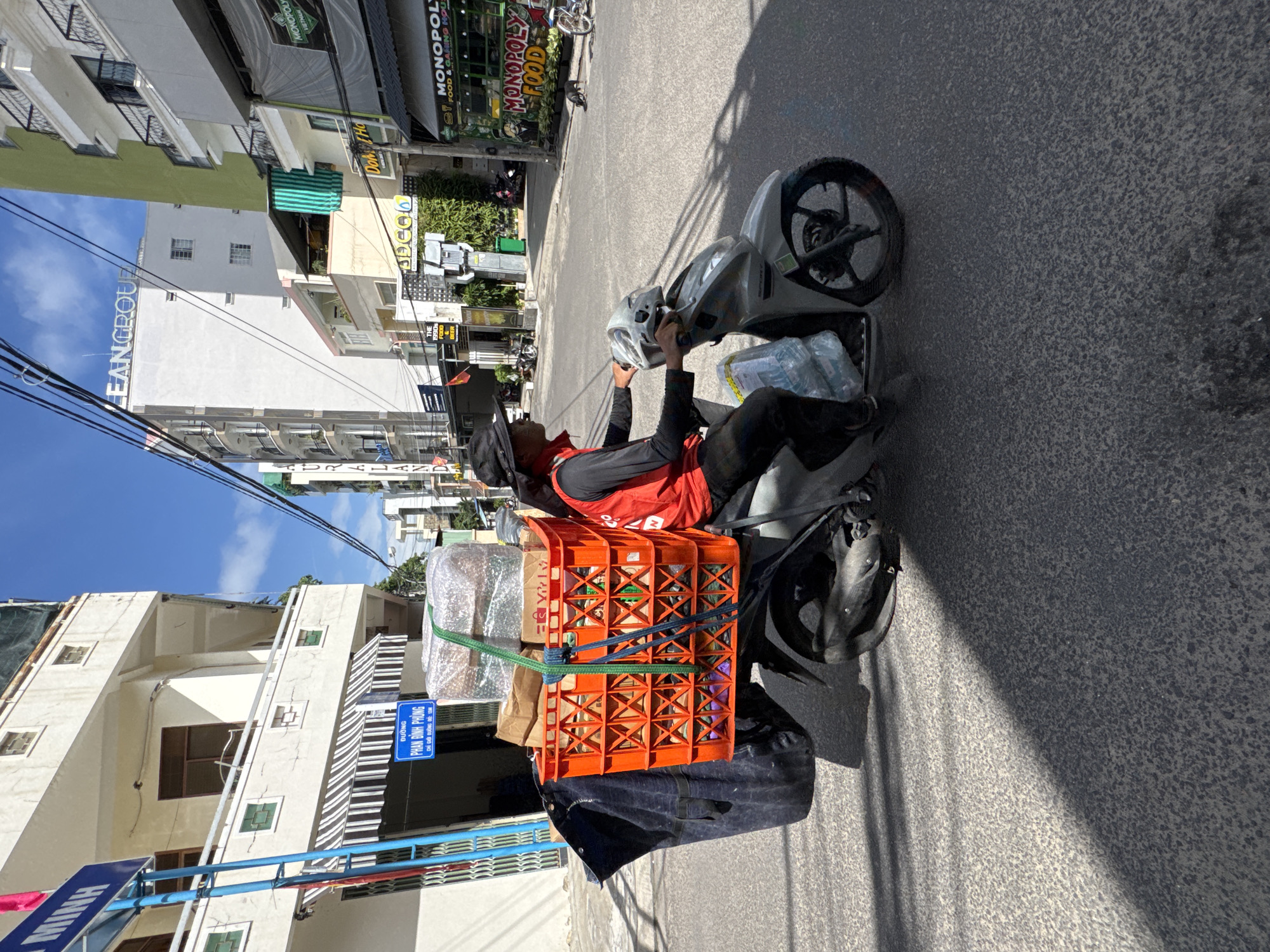 A man wearing a black hat and a red vest over a black long-sleeved shirt rides a silver scooter on a paved street in Nha Trang, Vietnam. The scooter has an orange plastic crate filled with items, including boxes wrapped in plastic, strapped to the back. A large dark blue bag is attached to the rear of the scooter. Buildings line the street, with awnings and signs visible. One sign reads "PHAN DINH PHONG" and another sign on a building says "LEAN GROUP". The sky is blue with some clouds. The sun casts shadows on the street. The overall scene depicts a delivery person navigating a city street.
