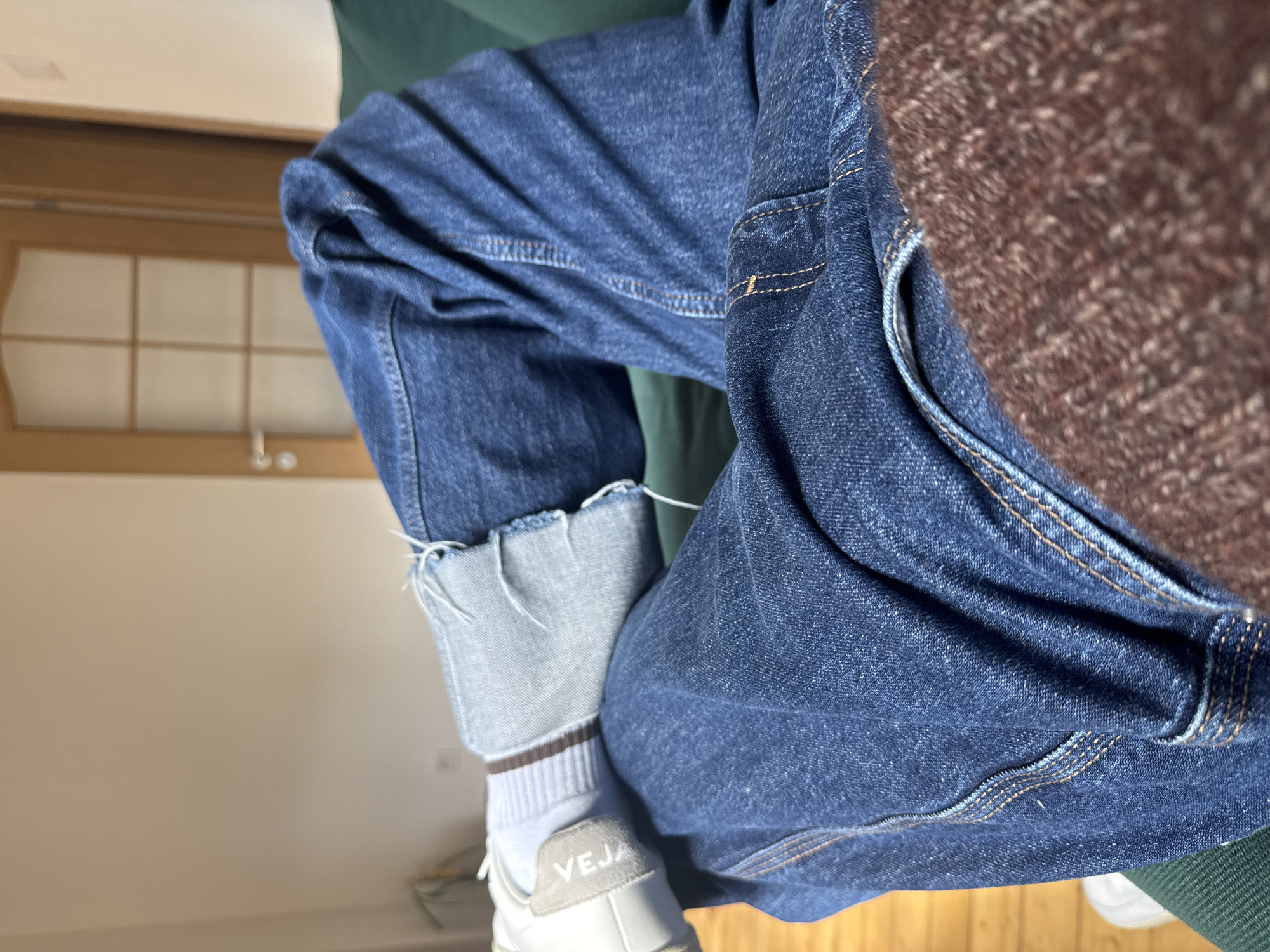 An indoor scene in Sofia, Bulgaria, depicts a first-person perspective view of a seated individual's lower body. The subject is wearing dark blue denim jeans with visible light brown stitching and some distressing/tearing near the right ankle, revealing lighter blue denim and loose white threads. A white ribbed sock with a thin brown stripe is visible on the left foot, paired with a white athletic shoe featuring "VEJA" on its heel tab. The individual's legs are crossed, with the right leg resting over the left, on a dark green upholstered surface, likely a couch or chair. A textured, brown, possibly knitted fabric covers the lower right foreground, obscuring part of the jeans. The background features a light beige wall. To the upper right, a light brown wooden door with a frosted glass panel in a grid pattern and a silver handle is partially visible. A light wood floor is discernible on the far left. The overall environment suggests a domestic setting.