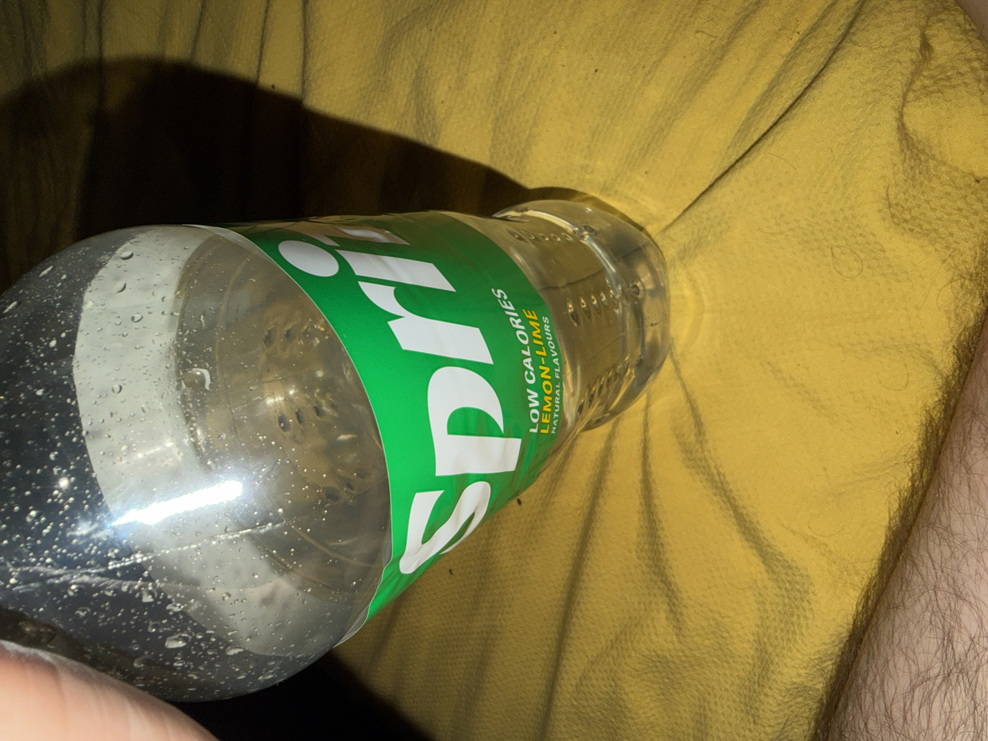 The image shows a partially full, plastic Sprite bottle lying on a crumpled yellow fabric surface, possibly a cushion or blanket. The bottle has condensation droplets on its exterior. Part of a person's arm with visible hair is visible on the right side of the frame. The location is not identifiable from the image.