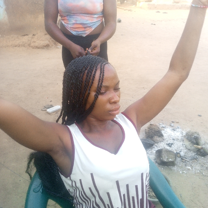 The scene is set outdoors in Ibreda, Nigeria. A dark-skinned female, seated in a dark green plastic chair, is having her hair braided by a second dark-skinned female standing directly behind her. The seated woman wears a white sleeveless V-neck top with brown vertical stripes on the lower portion and has numerous dark twist braids partially completed on her head. Her arms are raised and extended outwards, consistent with hair braiding. The standing woman wears a light-colored, tie-dye crop top and dark pants, with her hands engaged in the braiding process. The ground is composed of unpaved dirt or sand. To the right of the seated woman, an area of white ash and several dark rocks suggests the remains of a fire pit. Distant, blurred building structures are visible in the background.
