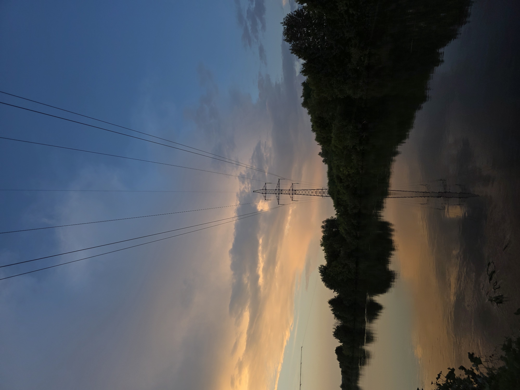 The image depicts a serene sunset over a river in Togashevo, Russia. The sky transitions from a soft orange and pink hue on the left, illuminated by the setting sun, to a deep blue on the right. Wispy clouds are scattered across the sky. A tall, metal electricity pylon stands prominently in the middle ground, with several power lines extending diagonally upwards towards the top of the frame. The pylon's reflection is visible in the dark, calm water of the river, which occupies the lower half of the image. Along the far bank of the river, a dense line of dark green trees forms a natural horizon. The overall atmosphere is peaceful and tranquil, characteristic of a rural landscape at dusk. There are no people or animals visible in the scene.