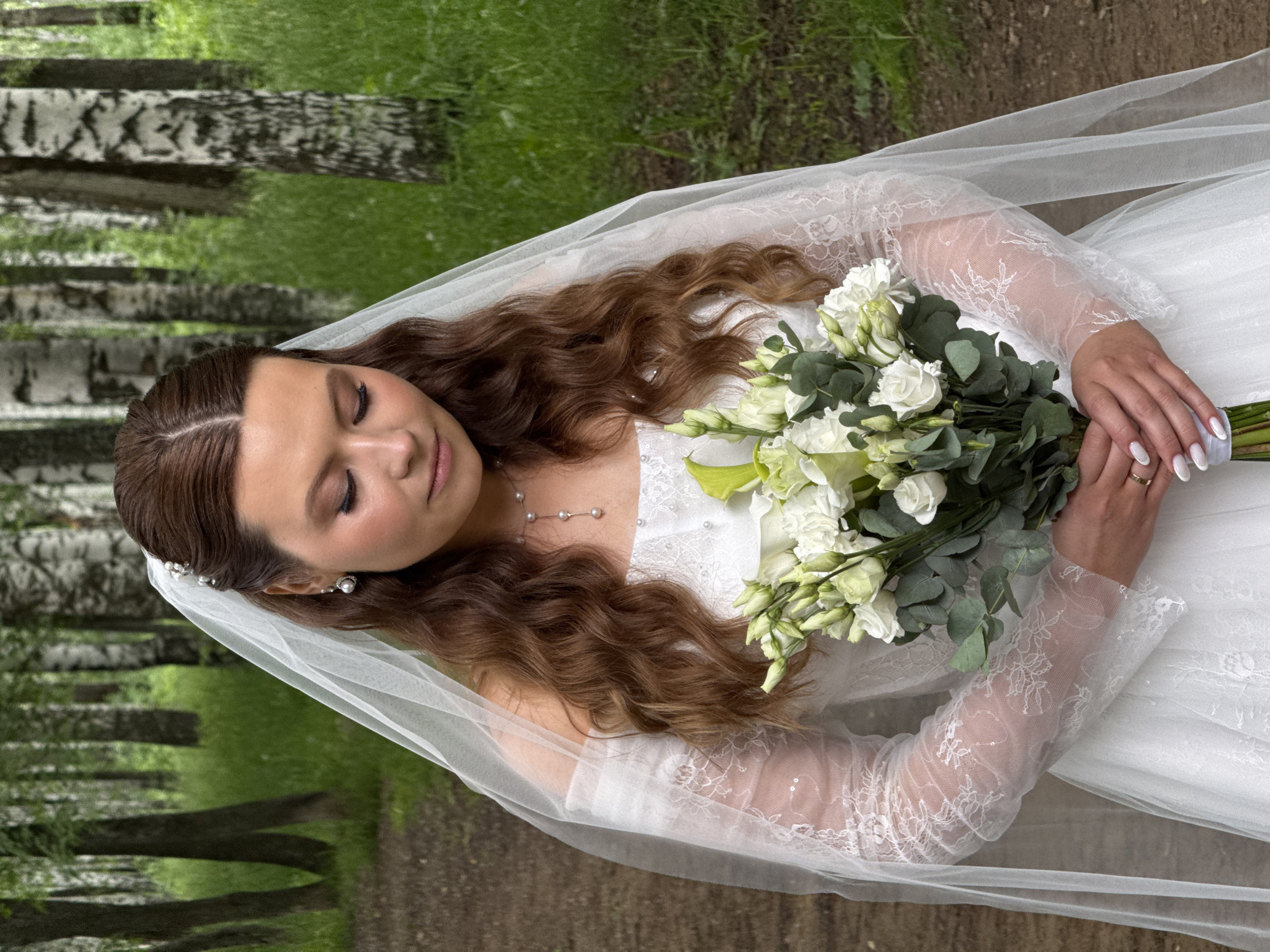 A medium shot, vertically oriented image depicts a serene bride in an outdoor, natural setting, likely a forest or park with prominent birch trees, given the location context of Chaikovsky, Russia. The bride is positioned diagonally across the frame, looking down with her eyes closed or cast downwards, conveying a peaceful and contemplative expression.
She has long, wavy, reddish-brown hair, styled with pearl hairpins, and wears a delicate, sheer white veil flowing behind her. Her bridal attire includes a long-sleeved white wedding dress featuring intricate lace patterns and possibly sequins or beads on the sleeves and bodice. A simple pearl necklace adorns her neck, and pearl earrings are visible.
Her hands, adorned with a white manicure and a wedding ring on her left ring finger, gently hold a voluminous bridal bouquet. The bouquet consists primarily of white flowers, possibly roses and lisianthus, interspersed with fresh green eucalyptus leaves and other foliage.
The background is softly blurred, showcasing a natural environment with a dirt path leading into a lush green area. Tall birch trees with their distinctive white and dark bark stand on either side of the path, receding into the distance, indicating a forest or woodland setting. The lighting suggests it is daytime, with soft, diffused natural light, implying an overcast or late afternoon sky. There is no visible text in the image.