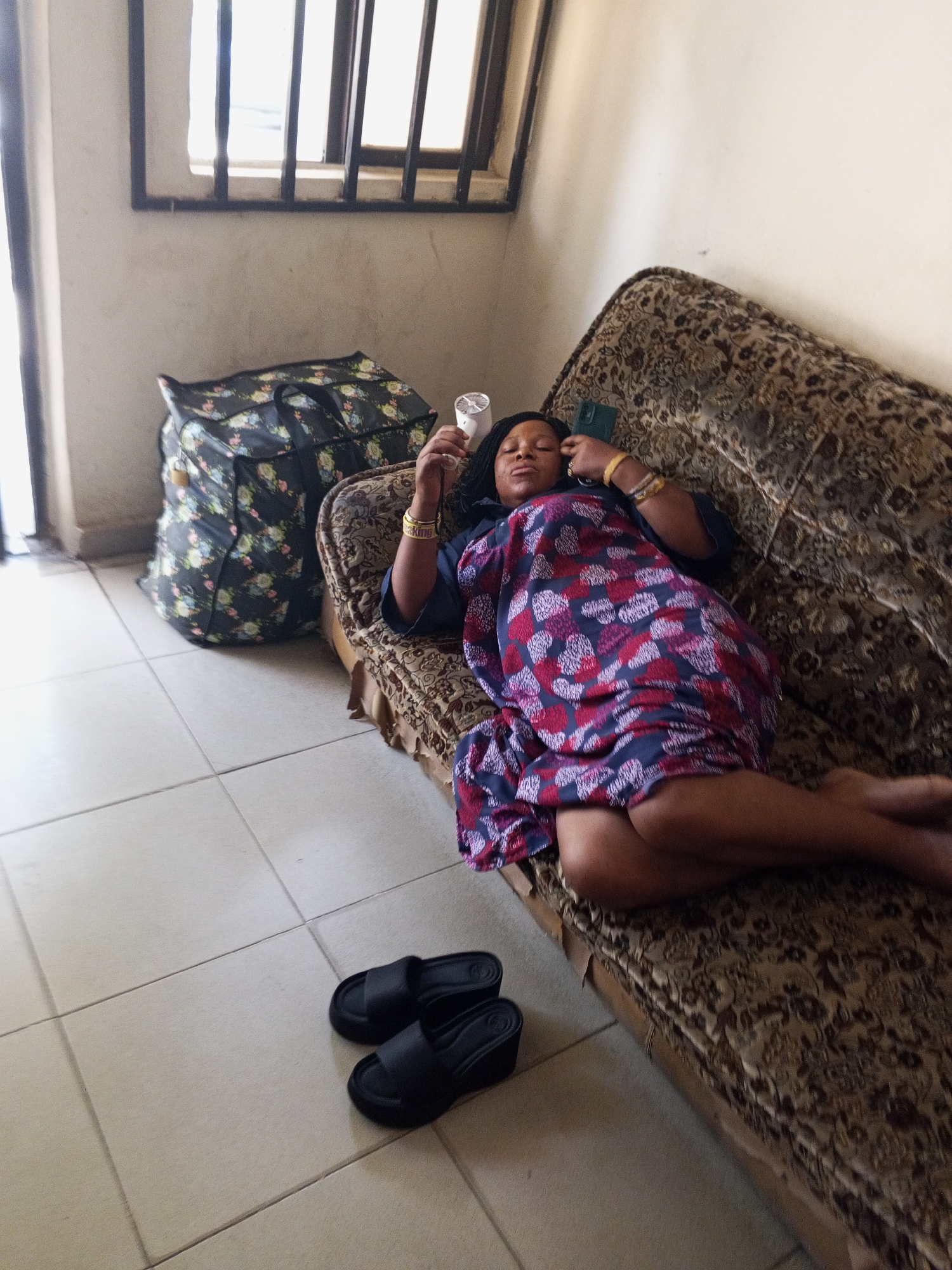 A woman is depicted lying on an old-fashioned patterned couch, seemingly resting or asleep, with her eyes closed. She is wearing a dark top and a colorful, patterned wrap or dress featuring red, blue, and white geometric or floral designs. On her right wrist, she wears several gold-colored bangles, one of which clearly displays the word "KING." A dark-colored smartphone is placed near her head, and she holds a small, white handheld fan in her left hand.
The couch, featuring a brown and beige floral or ornate pattern, appears well-worn. On the tiled floor in front of the couch, a pair of black slip-on slippers are neatly placed. To the left of the couch, a large, black carry-all bag with a vibrant floral pattern (roses and leaves) sits on the floor, secured with a small gold padlock.
In the background, a window with dark security bars allows bright natural light into the room, suggesting it is daytime. The walls are a light, neutral color, and the floor is made of large, light-colored tiles. The overall scene conveys a sense of quiet relaxation in an indoor setting.