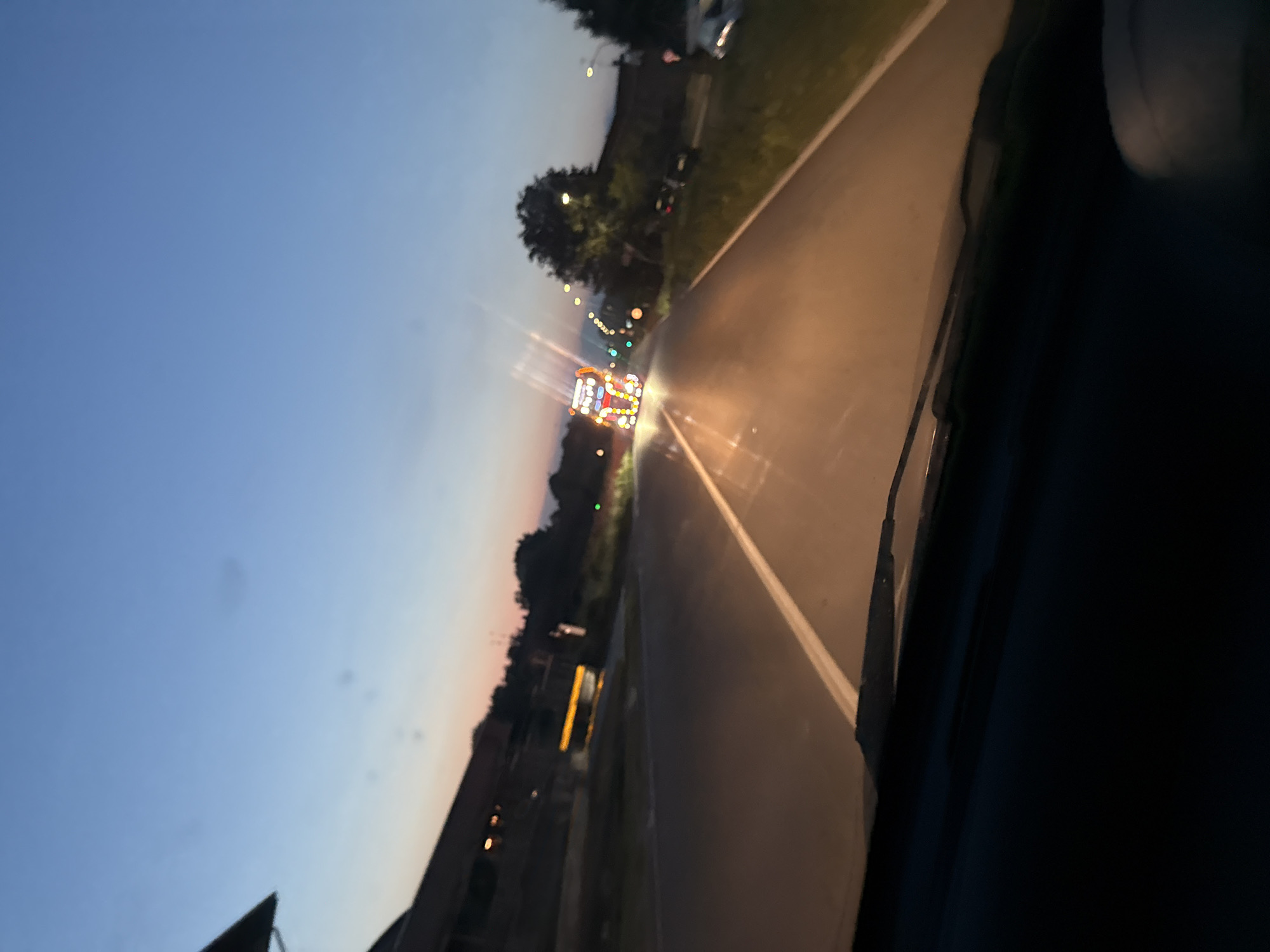 An outdoor, eye-level, slightly tilted shot captures a road at dusk, with a truck decorated with numerous lights driving away from the camera. The truck, positioned in the middle of the road, is illuminated by the colorful lights decorating its trailer and cab. The sky is a gradient of deep blue and orange hues, indicating twilight. The road, paved and with a white dividing line, curves gently to the right. To the left of the road, a building with lights visible in its windows is partially obscured by darkness and a roofline. Trees line the right side of the road, and streetlights are visible along the route. The foreground shows the dark interior of a vehicle, likely the one from which the photo was taken, with a windshield wiper visible. The overall scene is devoid of people. The location is Mezzana Sant'Antonio, Italy.