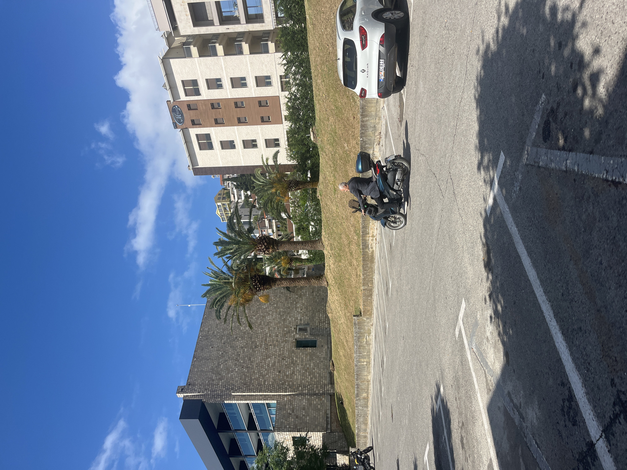 The image captures an outdoor scene, likely a parking area, under bright daylight with a clear blue sky and scattered white clouds. The setting appears to be an urban or suburban environment, possibly in Budva, Montenegro, as indicated by the location context and the Podgorica (PG) prefix on a visible license plate.
In the foreground and midground, an elderly person with grey hair is seated on a dark-colored scooter in a paved parking lot. The person is wearing a dark jacket and a lighter shirt. White parking lines are visible on the asphalt, along with shadows of trees cast across the ground, suggesting a sunny day. A white Renault Kadjar car is parked further back in the lot; its license plate "PG KC811" is clearly visible, with "KADJAR" written on its rear.
Behind the parking lot, a grassy slope is populated with several tall palm trees. Beyond the slope, there are multiple buildings. One prominent building features a light-colored facade with brown sections and numerous windows. A sign with the word "Melia" is visible on this building. Another building, partially visible to the left, has a dark brick exterior and modern glass windows. The architecture suggests a residential or commercial complex. No specific activity beyond parking and a person sitting on a scooter is evident.