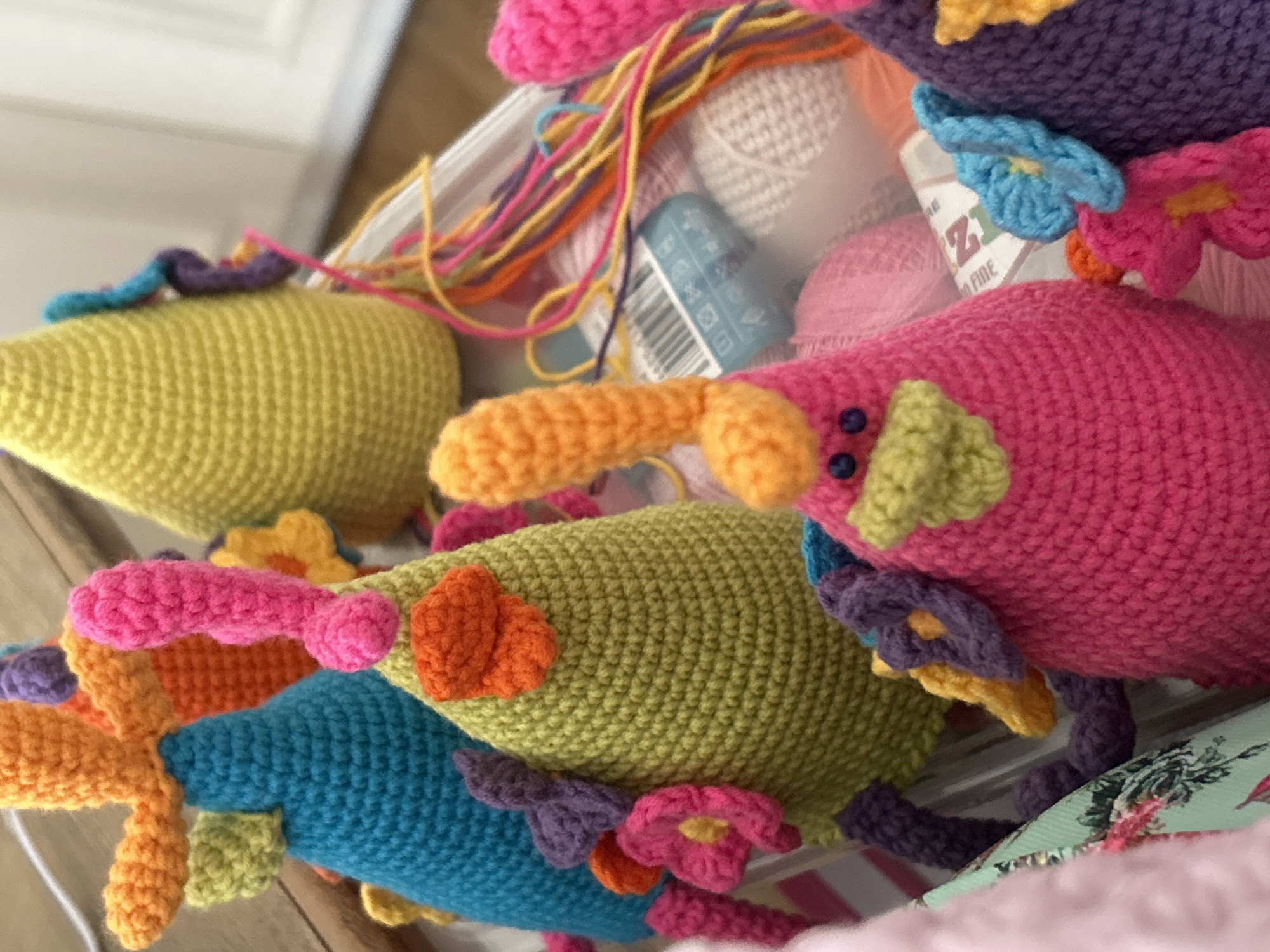 The image shows a close-up view of several colorful, crocheted toys arranged in a box or container. The toys appear to be abstract animal or creature forms, with various appendages, decorative flowers, and different yarn colors. The background includes spools and strands of yarn. The toys are piled together, suggesting they are stored or perhaps being prepared for sale. The toys feature different textures and patterns characteristic of crochet work. There is no indication that this image was taken in Plovdiv, Bulgaria, nor is there any information provided to indicate the location of the scene.