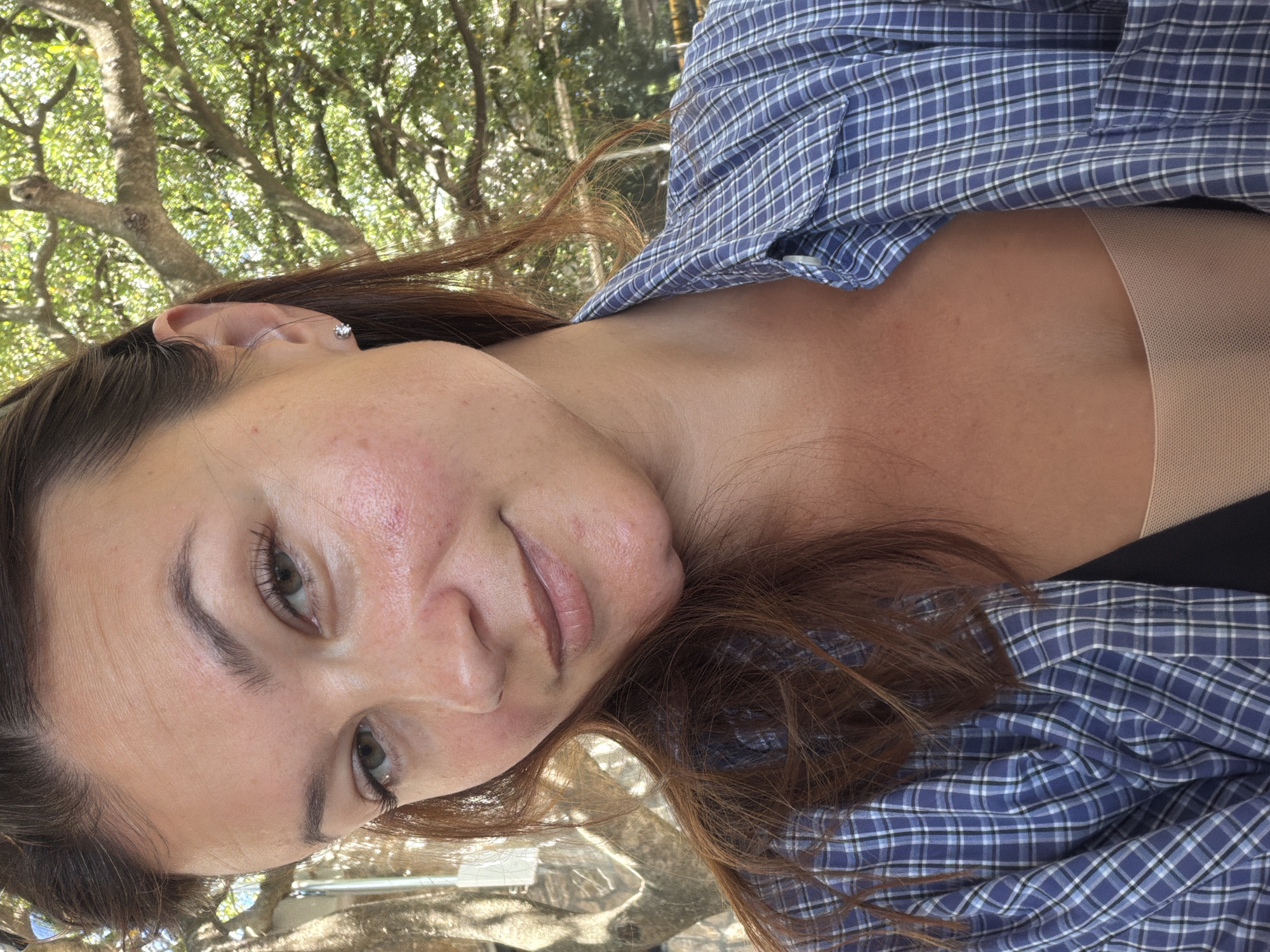 The image is a close-up, selfie-style portrait of a woman in Prijevor, Montenegro. She has medium-length brown hair, and she is looking at the camera with a slight smile. She is wearing a blue and white plaid button-down shirt over a tan-colored top. The background shows lush green trees and sunlight filtering through the leaves. The woman has a small stud earring in her visible ear. The overall lighting suggests an outdoor setting, likely during the daytime.