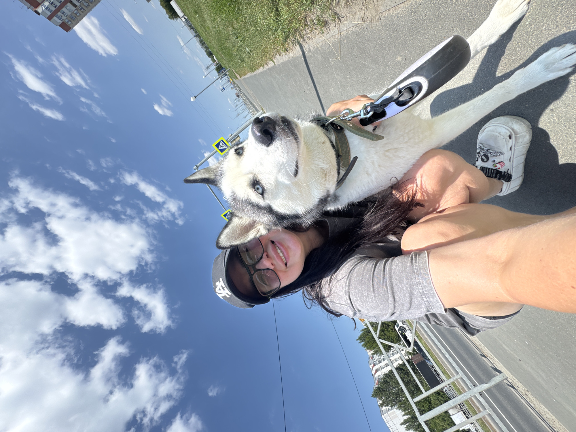 In a wide, low-angle selfie taken outdoors on a sunny day, a young woman with long dark hair, wearing glasses, a grey t-shirt, and a black baseball cap, smiles at the camera, her arm around her husky dog. The dog, with striking blue eyes and black and white fur, looks to its right. They are on a paved walkway next to a grassy area and a road. Behind them, a multi-lane road is visible with some vehicles, including a white truck and a dark-colored truck. In the background, apartment buildings and trees line the street, and streetlights and utility poles dot the landscape. The sky is bright blue with scattered white clouds. The image appears to have been taken in Tolyatti, Russia, suggested by the context of the prompt.