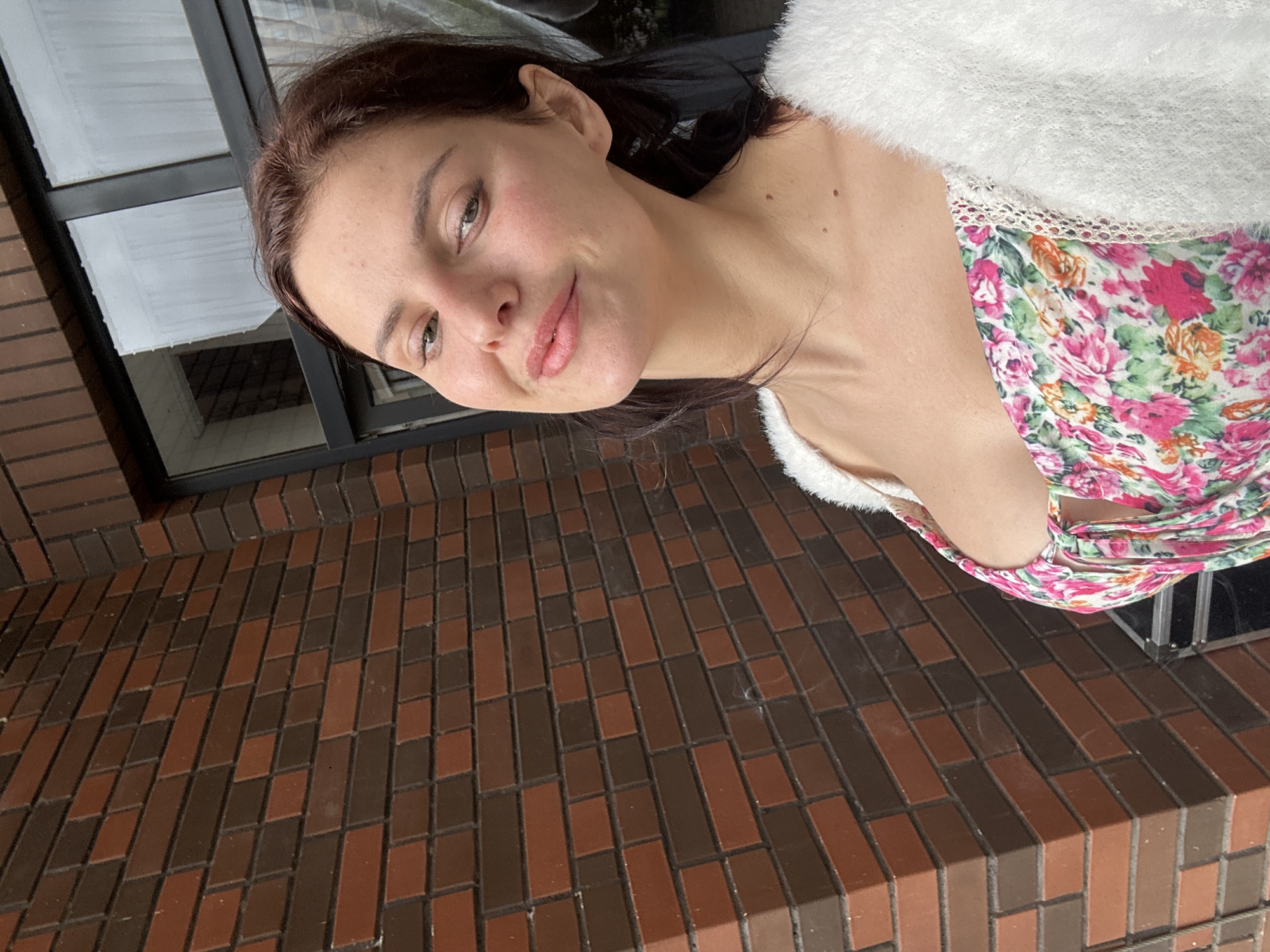 A woman with brown hair and light skin poses for a selfie outdoors. She is wearing a white, fluffy cardigan over a floral print dress with pink, green, and orange flowers. Her makeup includes pink lipstick and some blush. The background consists of a brown brick wall and a dark-framed window with a reflection of what appears to be white curtains. The scene is set in the city of Unknown, Unknown.