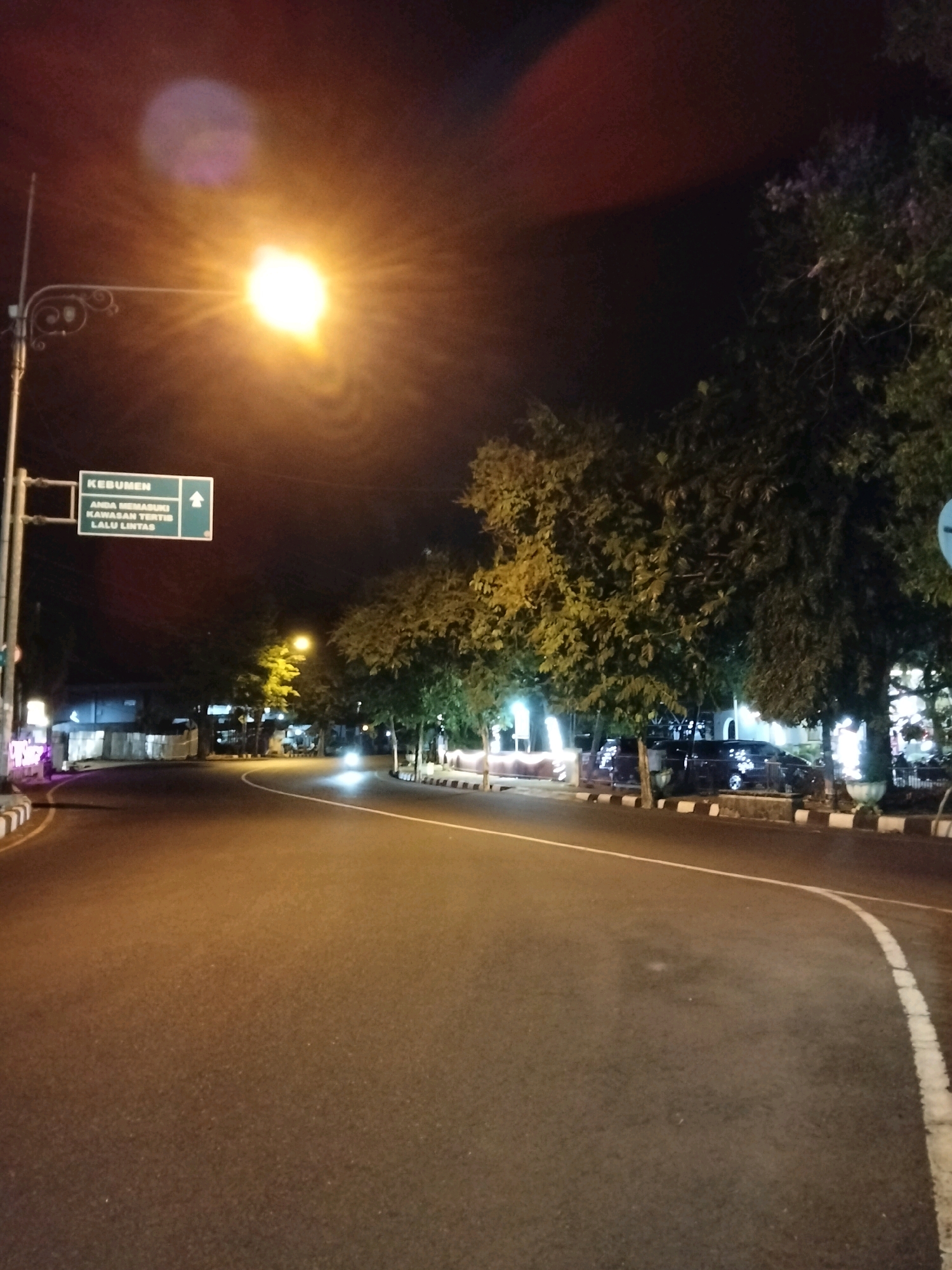 The image depicts a nighttime street scene in Purworejo, Indonesia, specifically on a road leading to Kebumen. A bright street lamp illuminates the area, casting a strong glow and creating lens flare effects in the upper portion of the frame. To the left, a directional signpost indicates "KEBUMEN" and "ANDA MEMASUKI KAWASAN TERTIB LALU LINTAS" (You are entering an orderly traffic area), with an upward-pointing arrow.
The road itself is paved asphalt with white lane markings. Trees line the right side of the road, their foliage dark against the night sky, with some street lights visible behind them. Several cars are parked or driving along the road. On the left side of the road, buildings and street lights suggest a commercial or residential area. The overall impression is one of an urban environment at night, with clear signage and traffic infrastructure. There are no visible people or specific actions occurring.