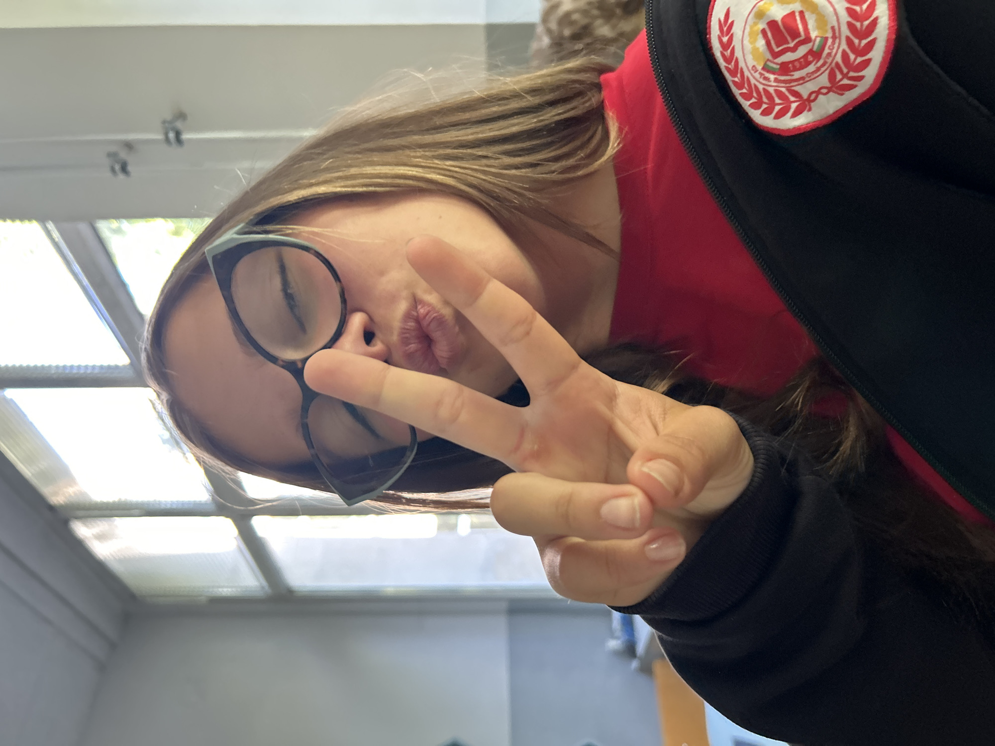 The media file is a close-up, rotated selfie or portrait, depicting a person, likely a young female, from the chest up. The individual has long, light brown hair and is wearing dark-framed glasses. Her head is tilted, and she is making a playful "duck face" expression with puckered lips. Her right hand is raised, making a peace sign with two fingers, which partially covers her face.
She is dressed in a red top or t-shirt, layered beneath a black zip-up jacket or hoodie. On the right shoulder of the black jacket, there is a circular patch. The patch features a white background with a red outline and a red laurel wreath design. Centrally placed within the wreath is an open book icon, and below it, the year "1974" is clearly visible. Below the date, partially obscured text in Cyrillic letters can be seen, which reads "СУ „Г. М. Димитров” Сливница", indicating a school named "G. M. Dimitrov" in Slivnitsa.
The background shows an indoor environment with a bright ceiling. Several rectangular skylights or large windows are visible on the ceiling, allowing abundant natural light to enter the space. This indicates the photo was taken during the daytime. The overall impression is one of casualness and youthfulness.