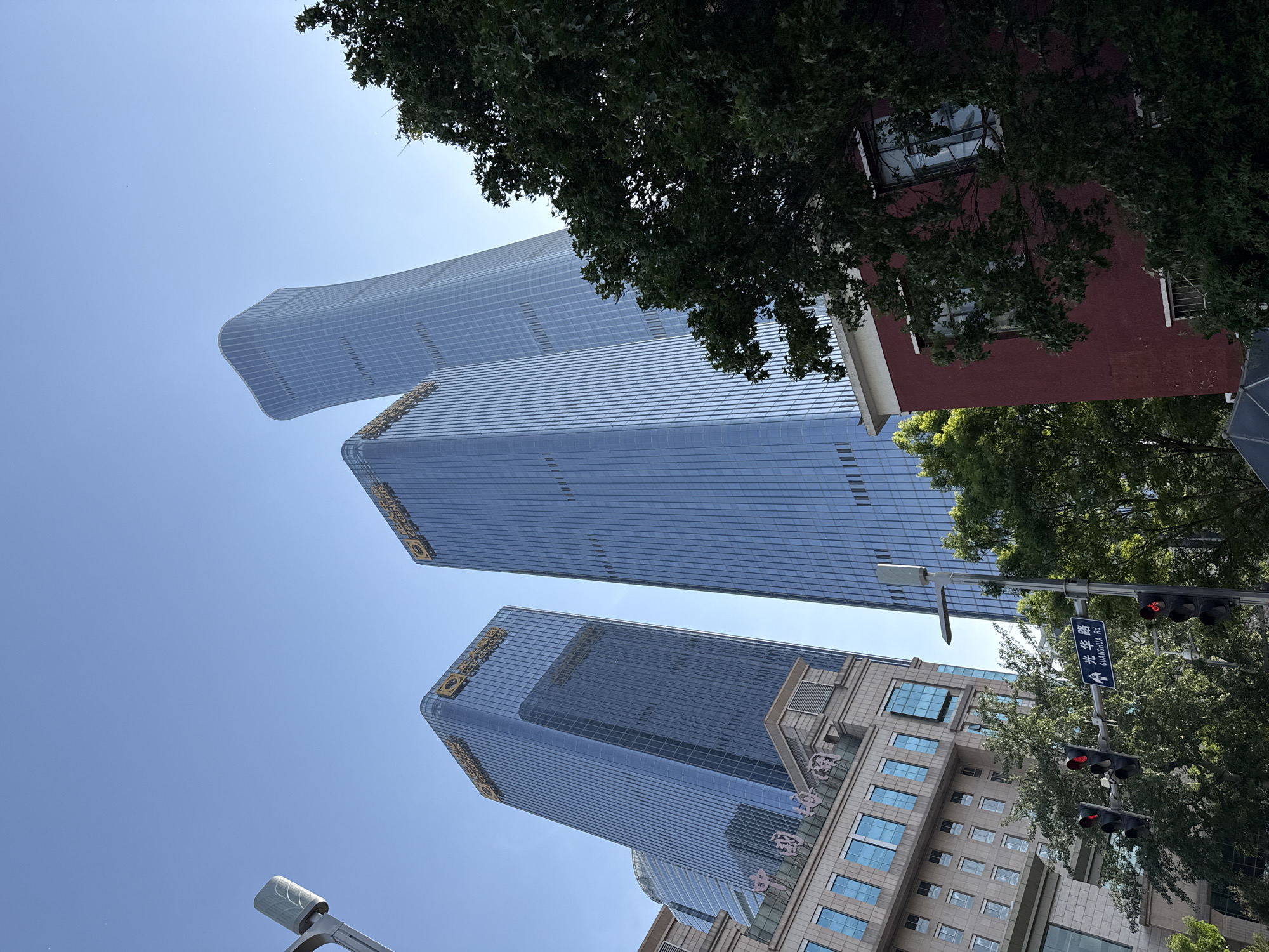 The image shows three tall, modern glass buildings in Beijing's Chaoyang District, China. The buildings, which appear to be commercial, ascend into a clear, blue sky. The lower building has a stone facade base. A street sign for Guanghua Road is visible along with traffic lights. A red-brick building and tree branches are in the foreground.