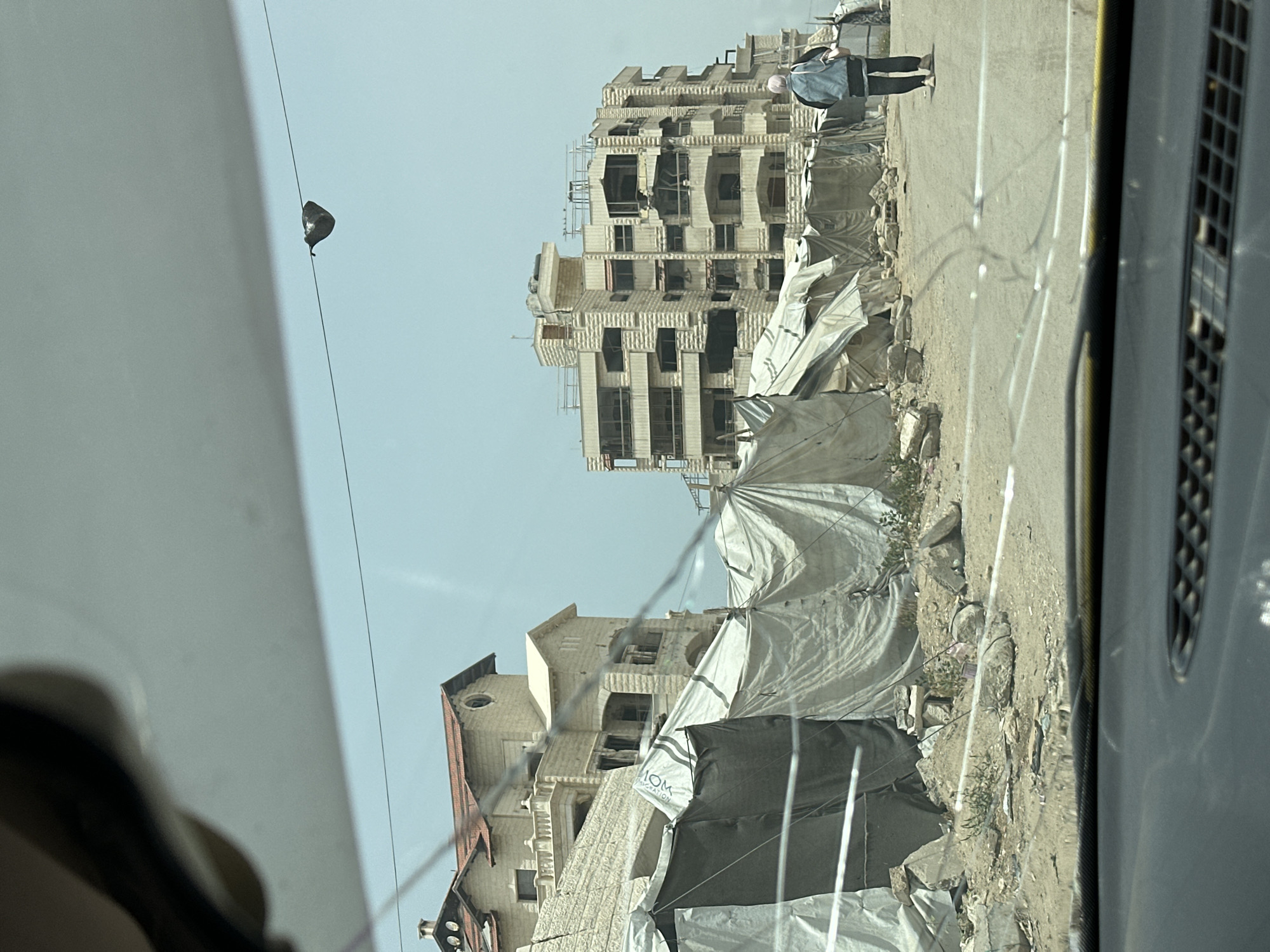 Captured from inside a vehicle, the image presents a street scene in what appears to be the city of Unknown, Unknown. Several partially constructed or damaged buildings stand alongside the road, some covered with white tarpaulin or other protective materials. Debris litters the roadside. One person is walking on the side of the road. A street light and wires cut across the upper portion of the frame. The vehicle's interior, including the dashboard and window, is visible in the foreground.
