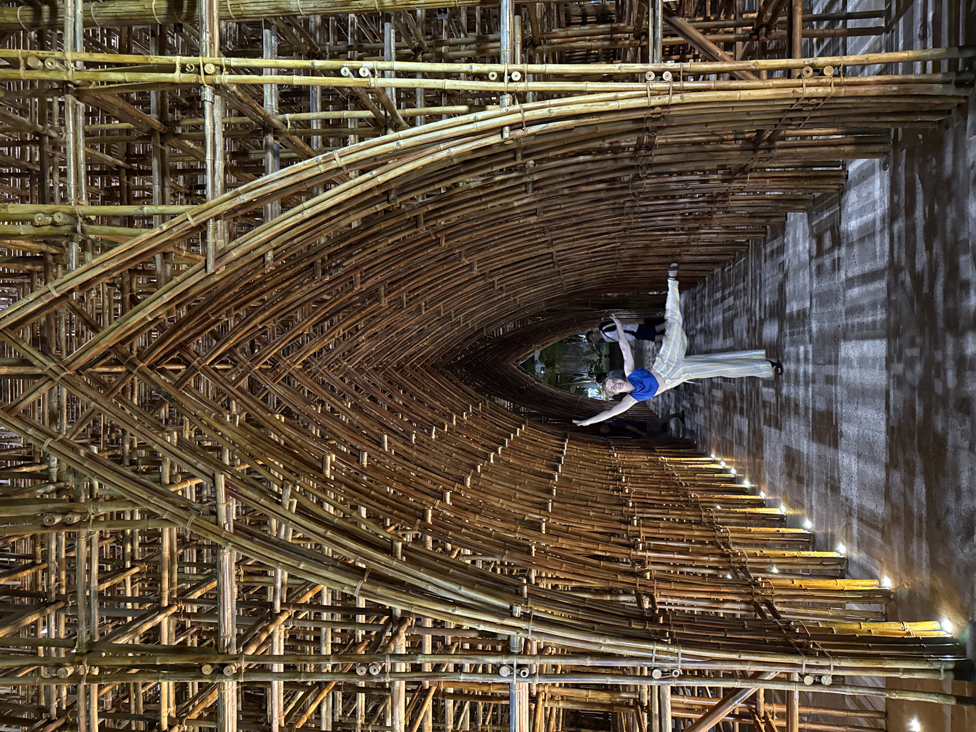 The image displays a person posing dynamically within a vast, intricately constructed architectural space made almost entirely of bamboo, located in Phú Quốc, Vietnam. The structure forms a towering, arched tunnel or vaulted canopy, with countless bamboo poles interwoven and secured to create a dense, organic lattice pattern that extends deep into the background.
In the mid-ground, on the right side of the vertically oriented image, a person is captured mid-pose. They are wearing a blue top and light-colored striped trousers, with their arms outstretched and one leg raised, suggesting a moment of balance or artistic expression. Their gaze is directed towards the camera, indicating they are intentionally posing.
The setting appears to be an indoor or covered environment, possibly an art installation, museum, or unique building hall. The ambient lighting is dim, contrasting with warm, artificial lights embedded along the base of the bamboo structure on the right side of the passage. These lights cast bright glows on the ground and highlight the texture of the bamboo, creating strong shadows on the adjacent wall. The dense, interwoven nature of the bamboo framework is a notable detail, showcasing a complex and robust construction technique. The scene evokes a sense of architectural wonder and intricate craftsmanship, likely captured during evening hours or in a controlled lighting environment. No visible text is present in the image.