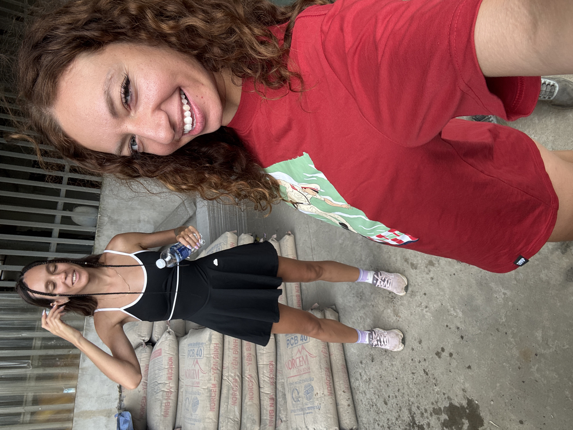 Two women are in Trung Tam Ward, Vietnam, standing in front of a row of stacked cement bags labeled "NORCEM YEN BINH PCB 40". One woman with curly hair is wearing a red T-shirt with a graphic on it, while the other is in a black dress with white trim, holding a water bottle. Both are smiling, and the scene is well-lit, suggesting an outdoor setting.