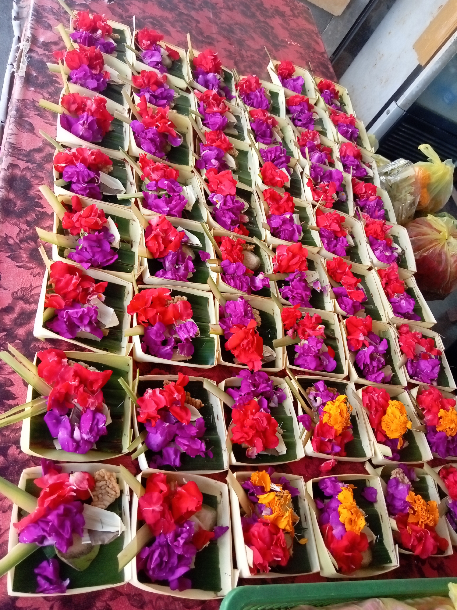 The image shows a large number of meticulously arranged offerings, likely for a religious ceremony, laid out on a patterned surface. The offerings are small, rectangular containers fashioned from palm leaves, each lined with a green leaf, and filled with a vibrant assortment of flowers in shades of red, pink, purple, and orange. Some offerings also contain small, irregular-shaped objects that appear to be dried grains or other natural materials. The offerings are arranged in neat rows and columns, creating a visually striking display. In the background, a yellow plastic bag and another bag with what appears to be produce are visible, suggesting a market or vendor setting. The lighting suggests an outdoor or well-lit indoor environment.