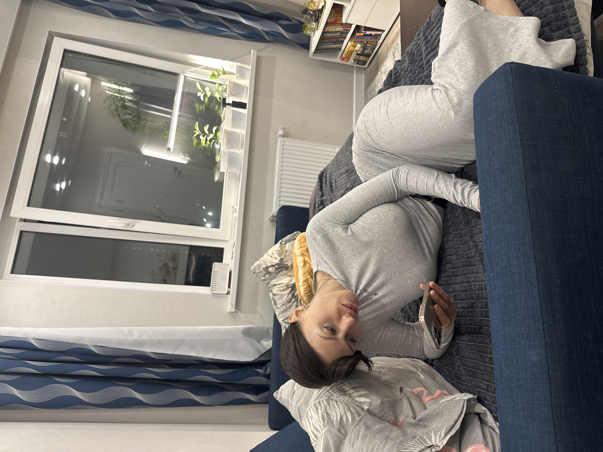 A woman is lying on a blue couch in a room. The room has a large window with a white frame, revealing an external view (not identifiable). There is a radiator under the window. The room also contains a white bookshelf with books. The woman is wearing gray clothing and holding a mobile phone. The scene is likely indoors, in a private residence. Location information (Murino, Russia) has not been visually confirmed.