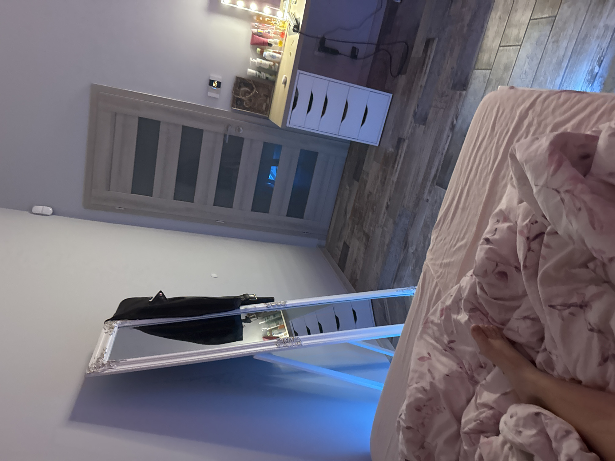 The image depicts an interior room, identified as being in Rezbartsi, Bulgaria. The scene is a bedroom featuring a bed in the foreground, a full-length mirror, a door, and a vanity station.
In the immediate foreground, a bed with a light pink duvet, adorned with a subtle floral or leafy pattern, occupies the lower left and central portion of the frame. A bare human foot is visible extending from under the rumpled duvet on the left side, indicating the presence of an individual in bed.
The floor is covered with distressed grey and brown wooden plank flooring.
On the left side of the room, a tall, ornate white-framed full-length mirror leans against a light grey wall. A strong blue light source, likely an LED strip, is positioned behind the mirror, casting a blue glow onto the wall and floor. The mirror's reflection shows a white five-drawer storage unit (typical of a vanity setup) laden with various cosmetic bottles and containers. A black garment, possibly a jacket or coat, is draped over the upper right corner of the mirror's frame.
Further into the room, centrally positioned, is a light-colored wooden door with multiple horizontal frosted glass panels. The door is closed, and a blue reflection from the mirror's light source is visible on its panels. A silver door handle is present. To the left of the door, high on the wall, a small white rectangular sensor or device is mounted. To the right of the door, another white digital display unit, possibly a thermostat, shows the number "26".
On the right side of the frame, a light wooden desk or vanity top is visible, supported by a white five-drawer unit similar to the one reflected in the mirror. The vanity surface holds numerous cosmetic and personal care products, including bottles, tubes, and a portion of an illuminated makeup mirror with visible bulbs. A black electrical cord and plug are present on the floor next to the drawer unit.
The room's walls are painted a uniform light grey. The primary light sources are the ambient room light, the blue LED behind the mirror, and the illuminated vanity mirror.