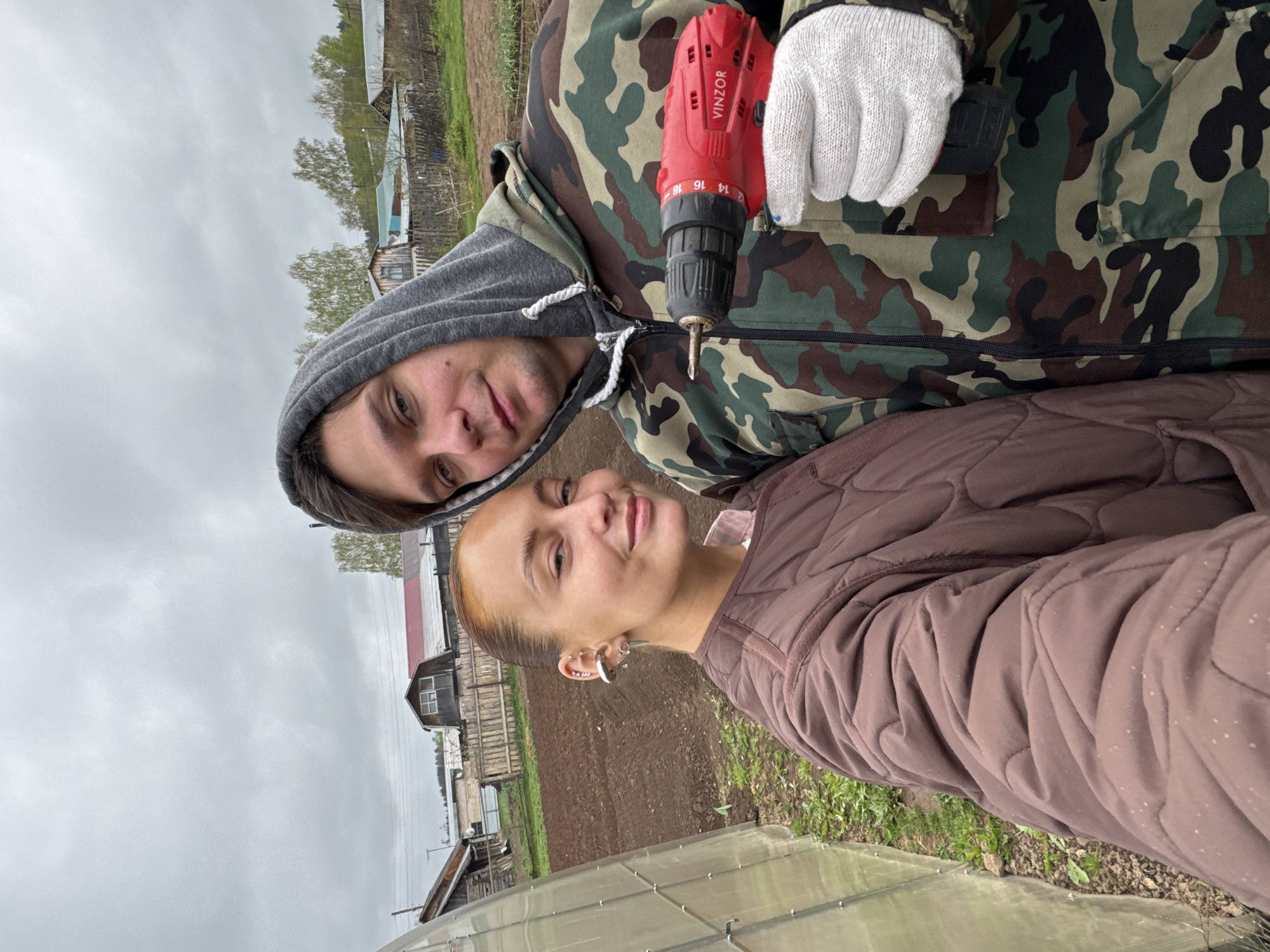 Two individuals are present. The individual on the top is male, wearing a camouflage hooded jacket, grey hoodie, and white glove, and is holding a red electric drill. The bottom individual is female, wearing a brown quilted jacket. The background shows an outdoor scene. There are buildings and tilled soil. The sky is overcast. The location is Chastyye, Russia.