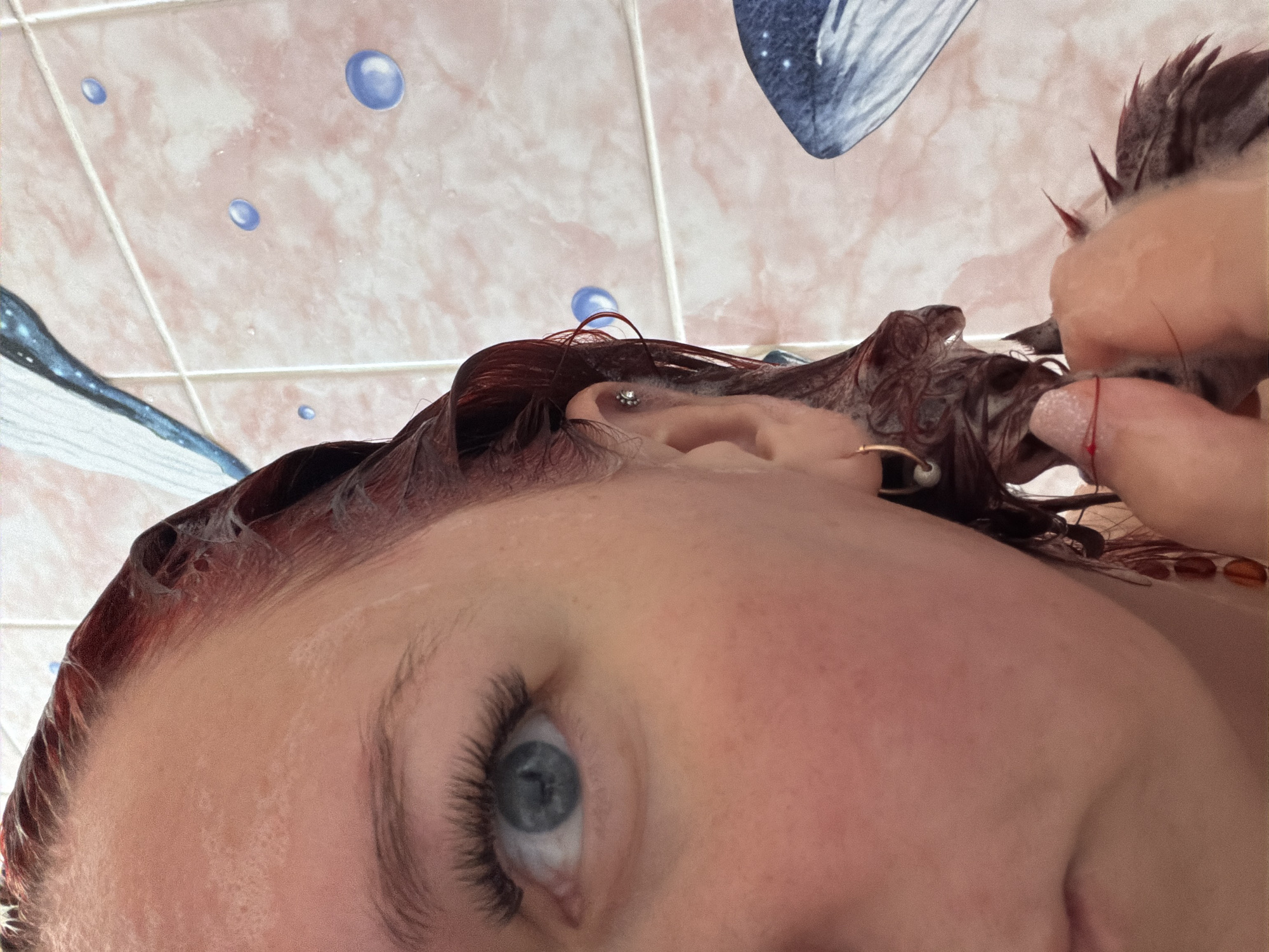 A close-up, angled shot captures a person with fair skin and light blue eyes. Their hair is dark red and appears wet, possibly freshly dyed. The person has multiple earrings on their left ear. The background reveals pink and white tiled walls with a pattern of blue bubbles and marine life. The image location or information concerning Chaikovsky, Russia, is not evident from the photograph. The main action seems to involve the person’s hair.