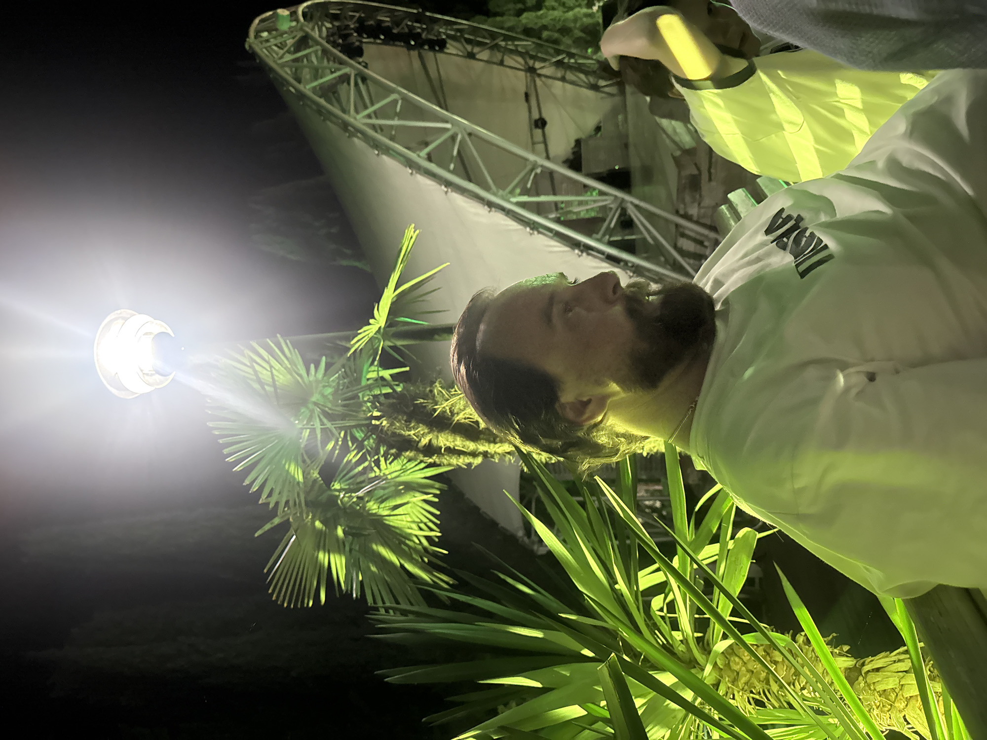 In the city of Yalta, Ukraine, at night, a man with a beard and dark hair is seated and looking to his right. He is wearing a white sweatshirt with black text on it. In the foreground, to the left, there are large, bright green leaves of a palm tree. Behind the man, there is a large, illuminated, curved metal structure, possibly a stage or canopy, with additional greenery visible in the background. A bright light is positioned above and to the left of the man, casting a strong glare. Another person, partially visible, is seated to the right of the man. The overall lighting suggests an outdoor evening event.